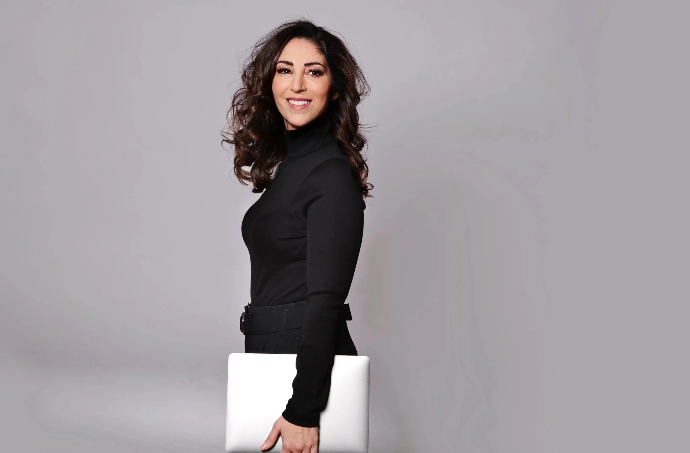 Eine Frau in schwarzem Pullover und schwarzen Hosen hält ein Laptop und lächelt vor einem grauen Hintergrund.