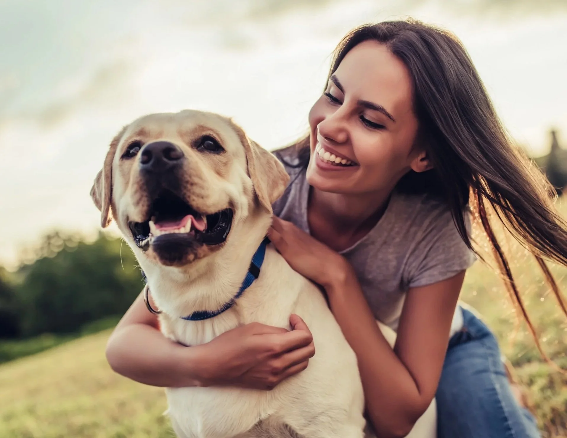 A woman smiling and cuddling a happy Labrador Retriever outdoors during daytime.