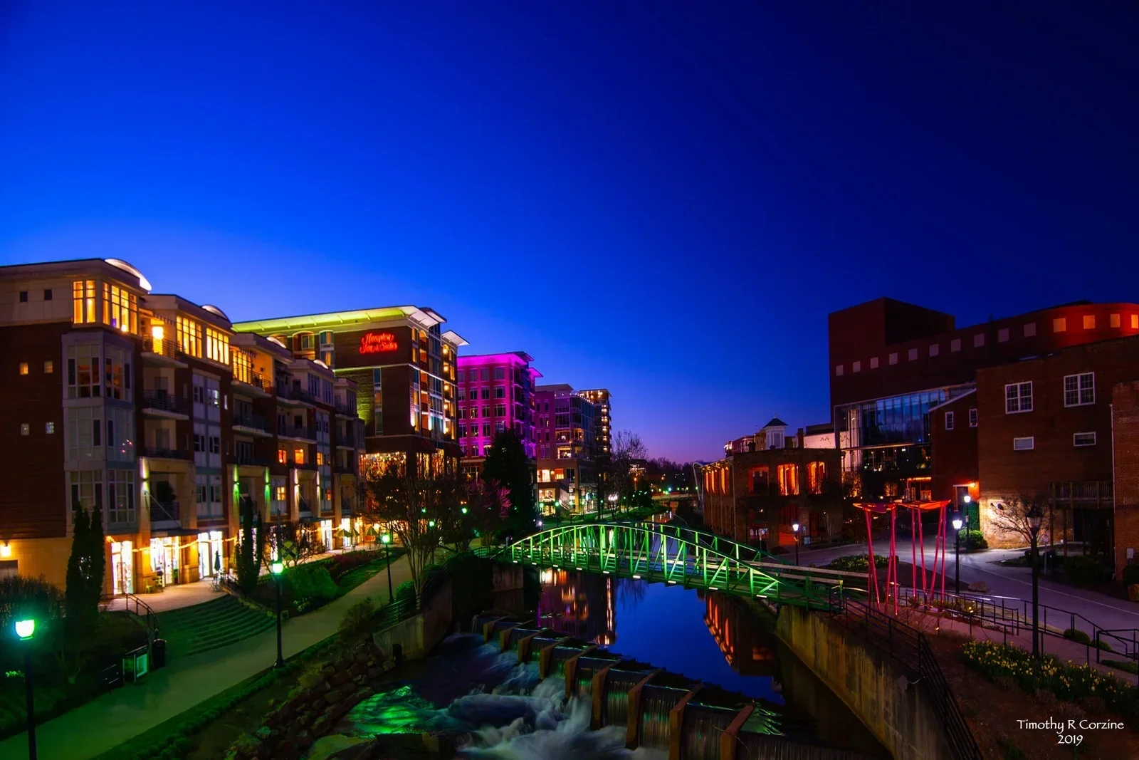 Night view of a modern cityscape with colorful illuminated buildings, a small river with a waterfall, a green-lit pedestrian bridge, and a vibrant sky.