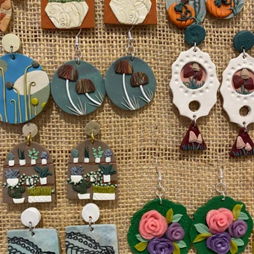 Various handmade earrings hanging on a textured backdrop, featuring mushrooms, floral shapes, and abstract designs in different colors.