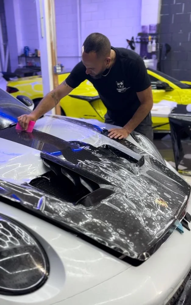 A man is cleaning the hood of a white sports car with water and a sponge inside a garage. Two other sports cars, one yellow and one purple, are visible in the background.