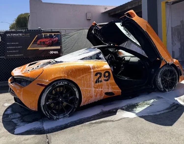 Orange sports car with scuffed paint, number 29, being washed and foamed with soap water in an outdoor car wash area.