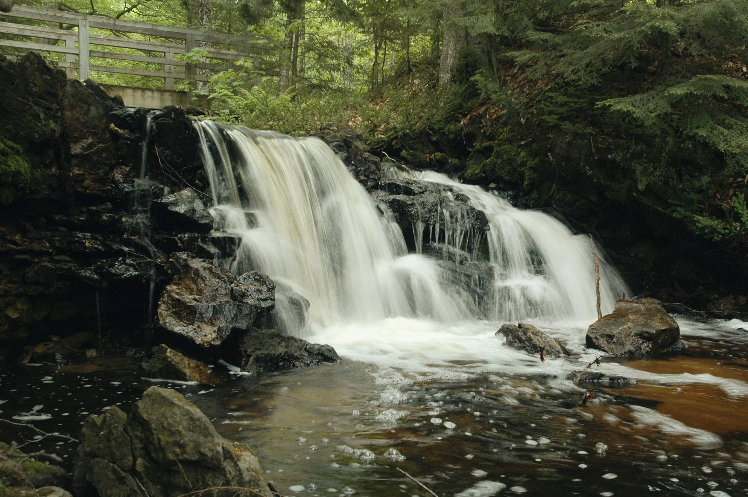 A small waterfall flowing over rocks in a forested area, with a wooden fence visible in the background.