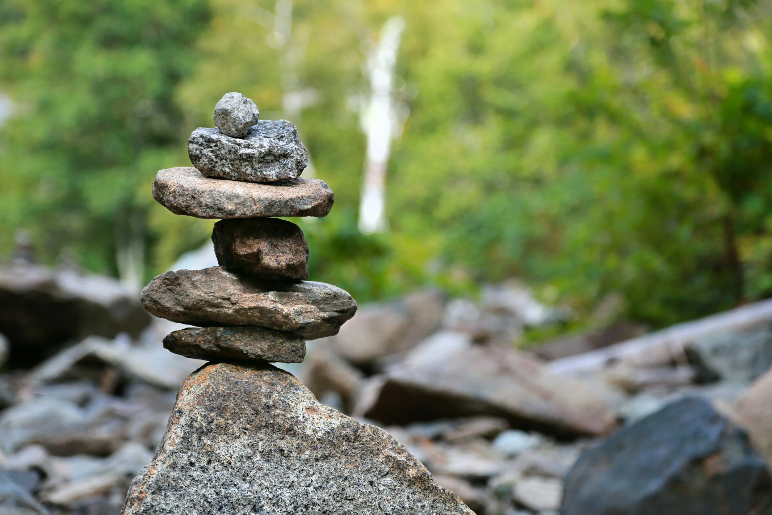 A stack of six balanced rocks on a large base rock in nature with blurred green trees in the background.