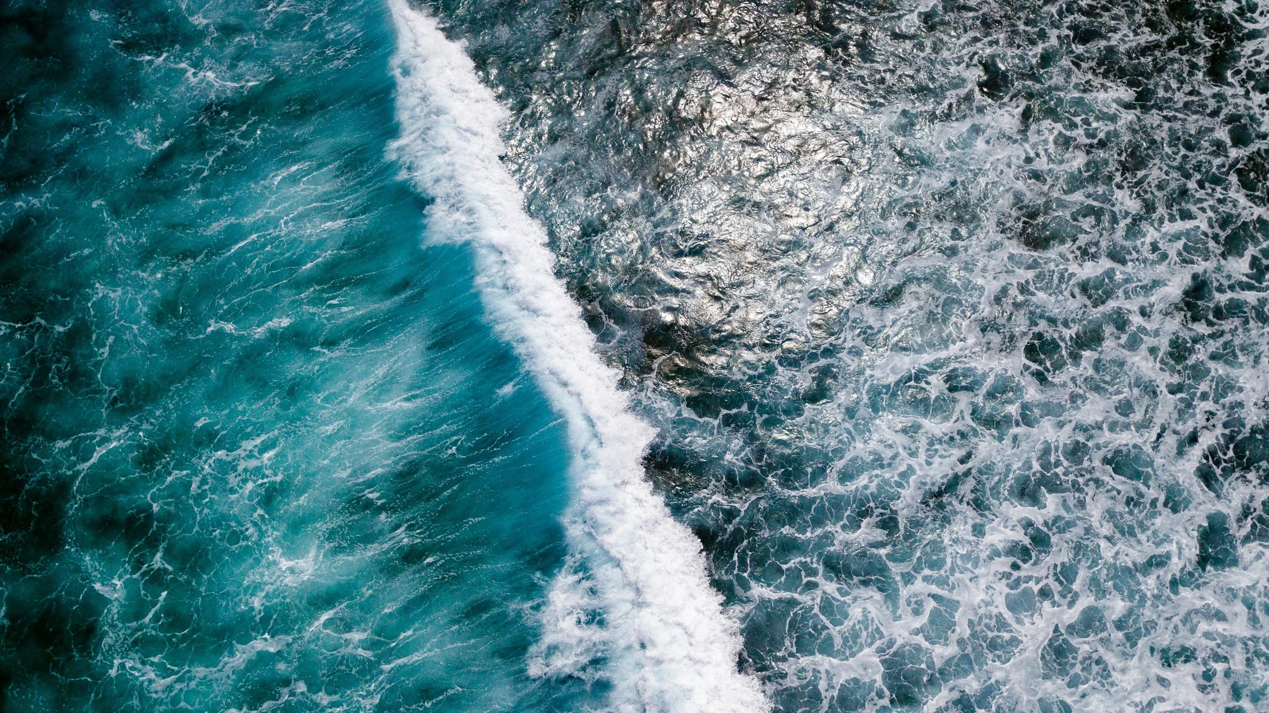 Aerial view of ocean waves crashing, showing white foam and deep blue water.