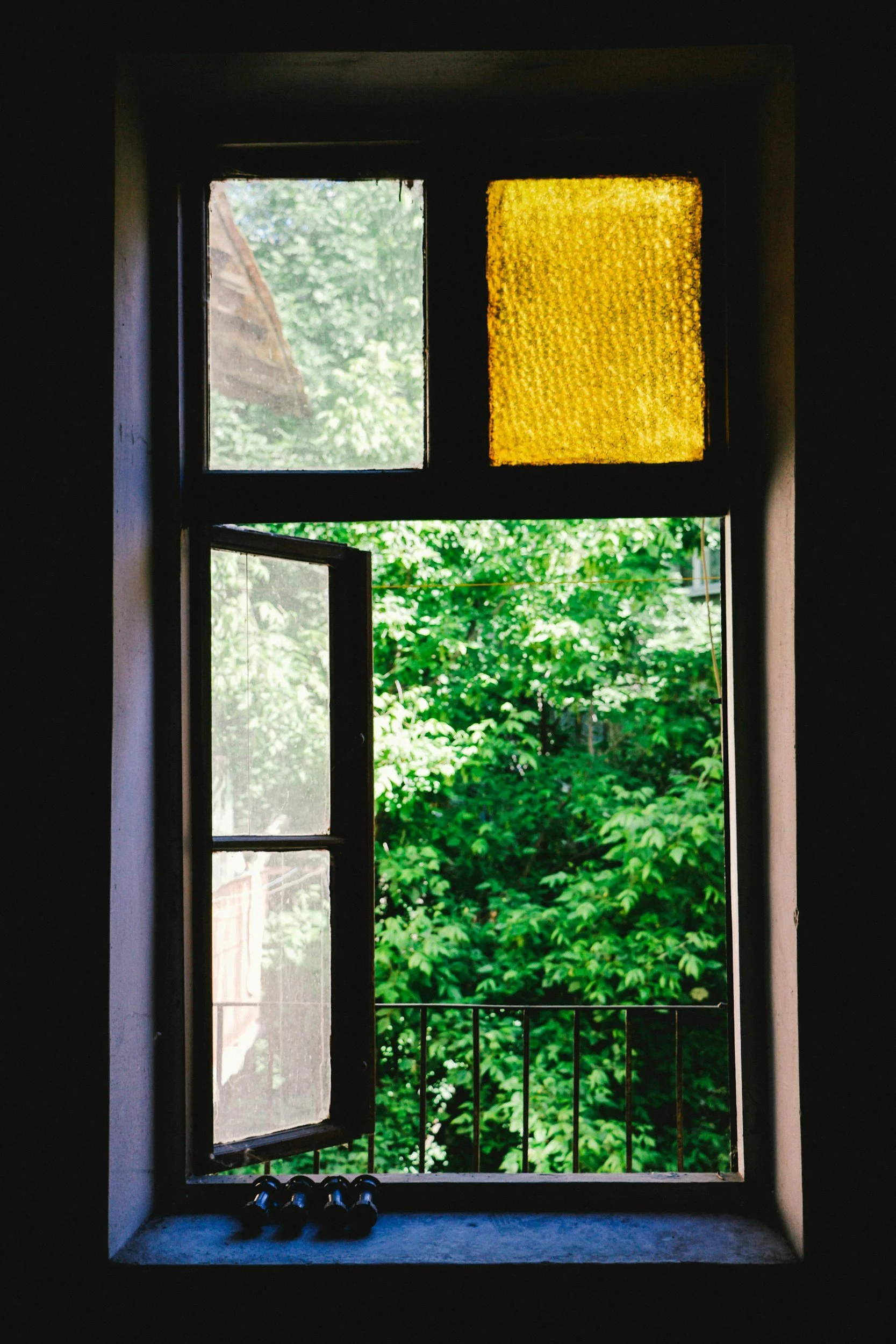 Open window with black frame and yellow stained glass window at the top, showing green leaves outside and a railing below the window.