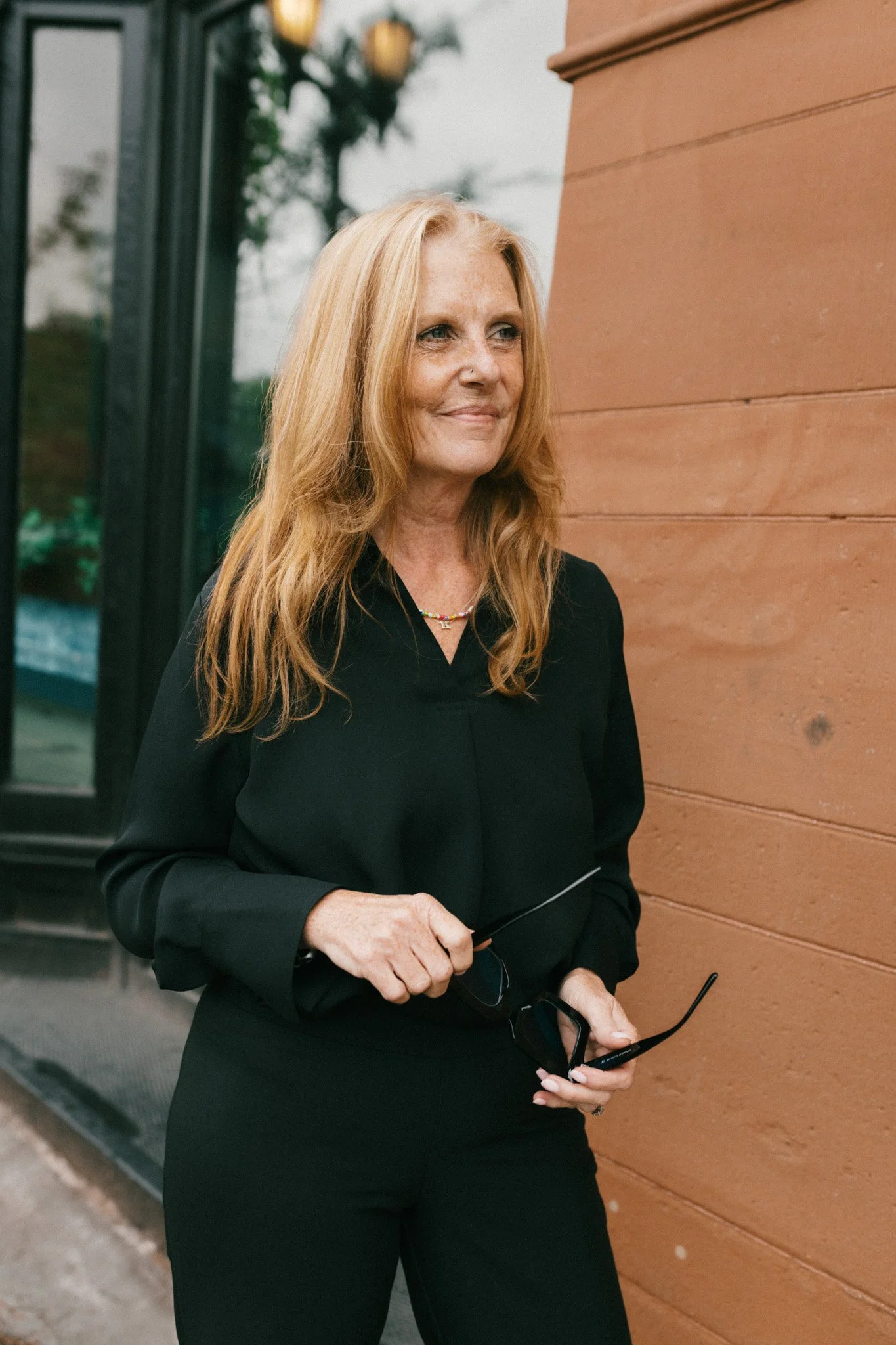 A woman with long red hair, wearing a black outfit and jewelry, standing outside near a wooden wall, holding glasses and a pair of sunglasses.