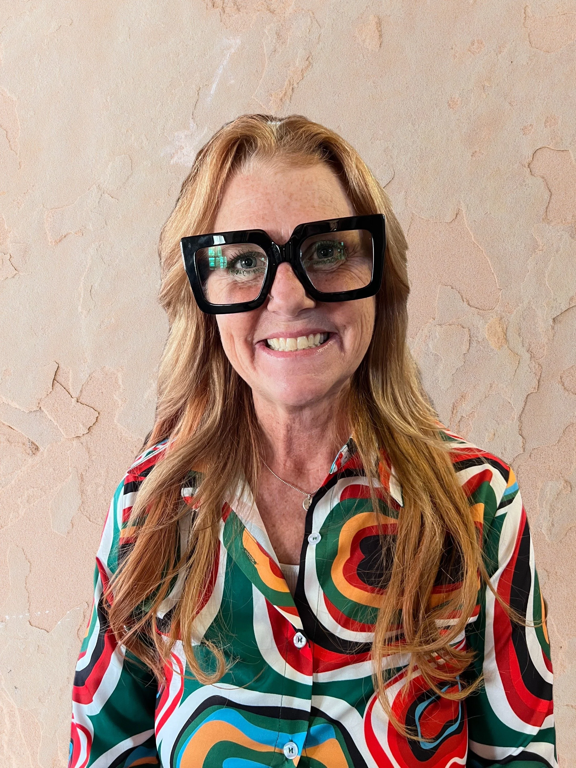 A woman with long red hair wearing large black glasses and a colorful, patterned shirt standing against a textured beige wall.