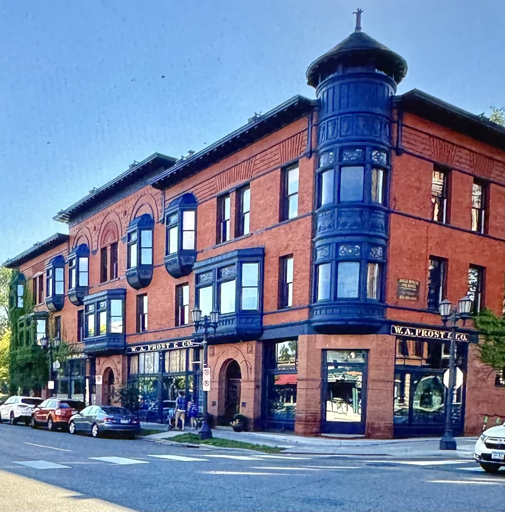 A historic brick building with blue window frames and a corner turret, located on a city street with parked cars and people walking.
