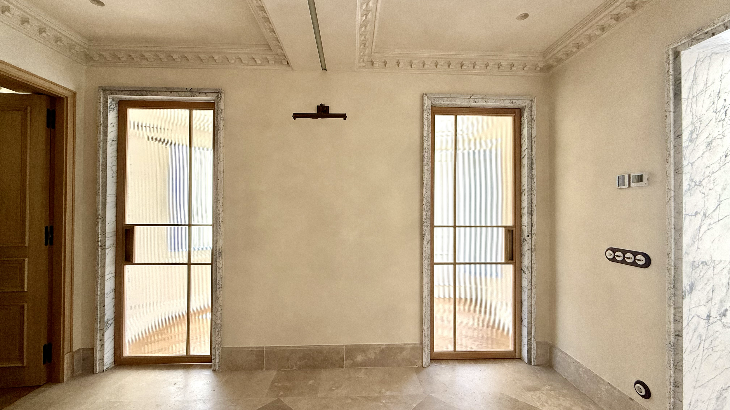 Empty room with two glass doors framed with white marble, beige walls, and a decorative ceiling trim.