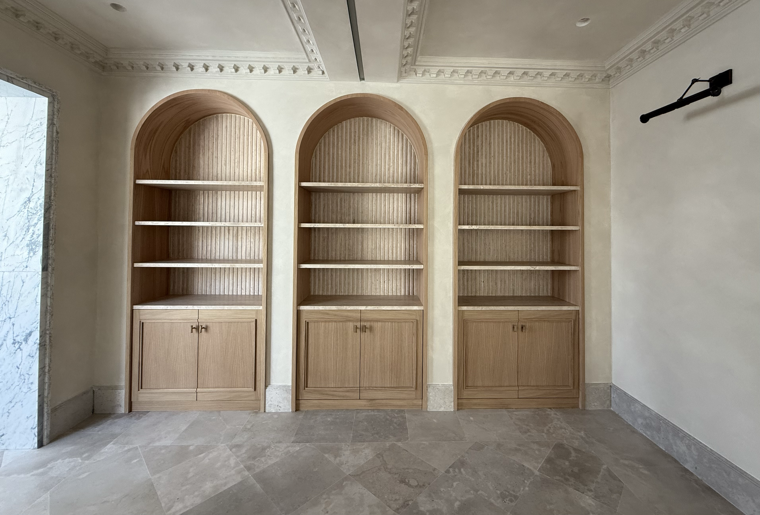 Three built-in wooden shelves with arched tops and lower cabinets in a beige room with decorative crown molding and tiled floor.