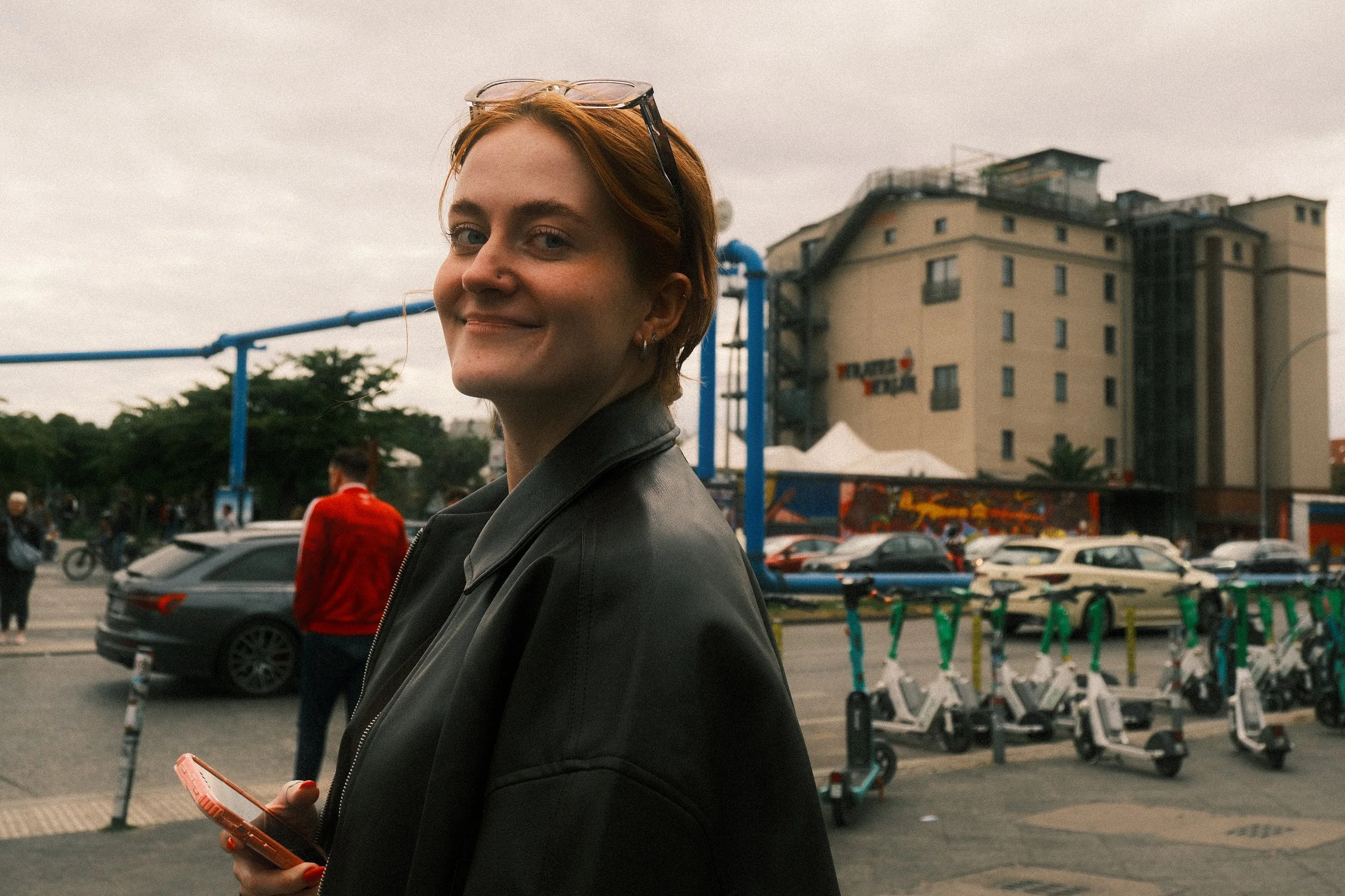 A woman with short red hair, wearing a black leather jacket, holding a phone, and smiling at the camera on a city street with cars, bicycles, and a multi-story building in the background.