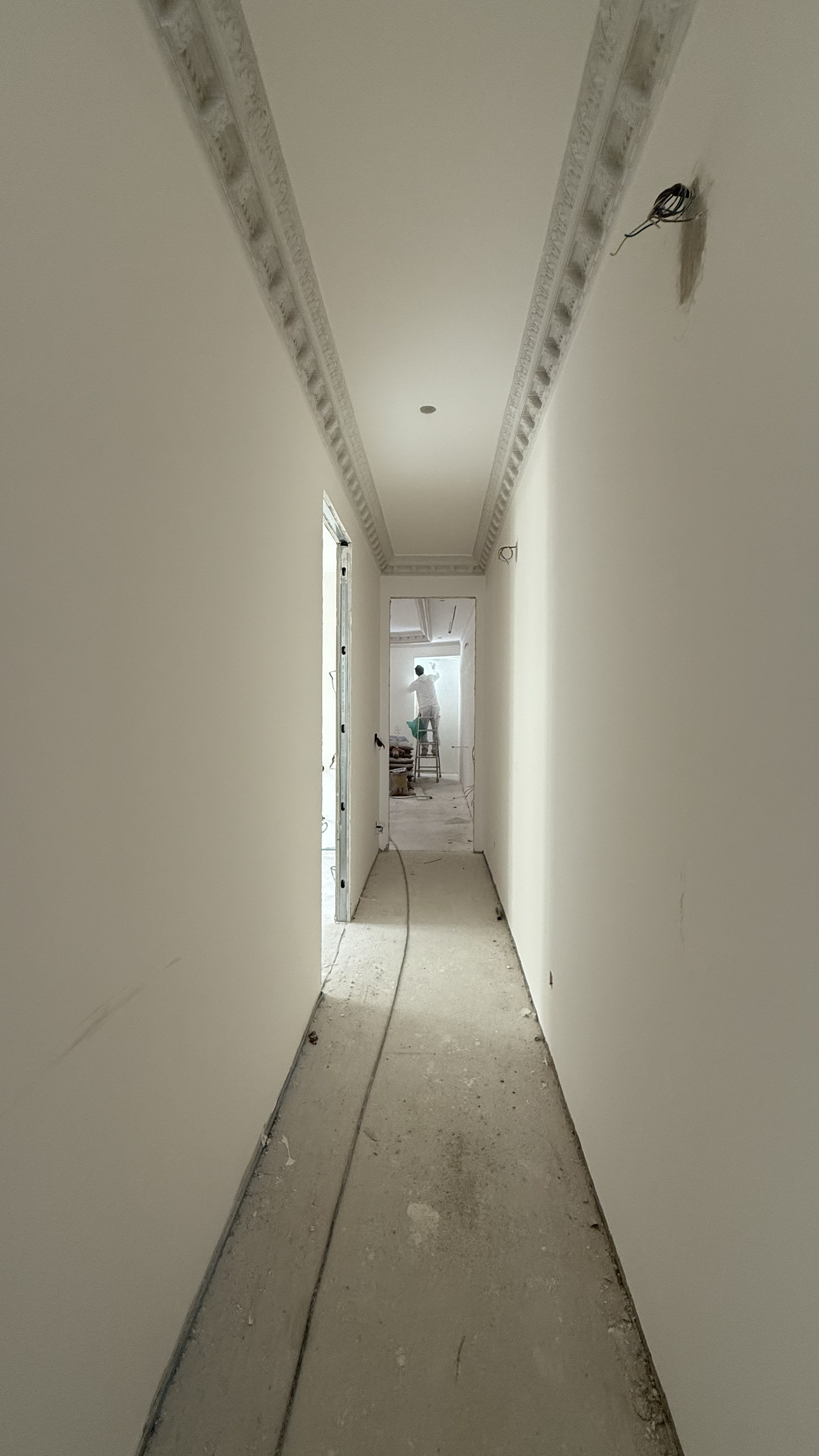A hallway under construction with unfinished flooring and wiring hanging from the ceiling. A person is working on a ladder in the background.