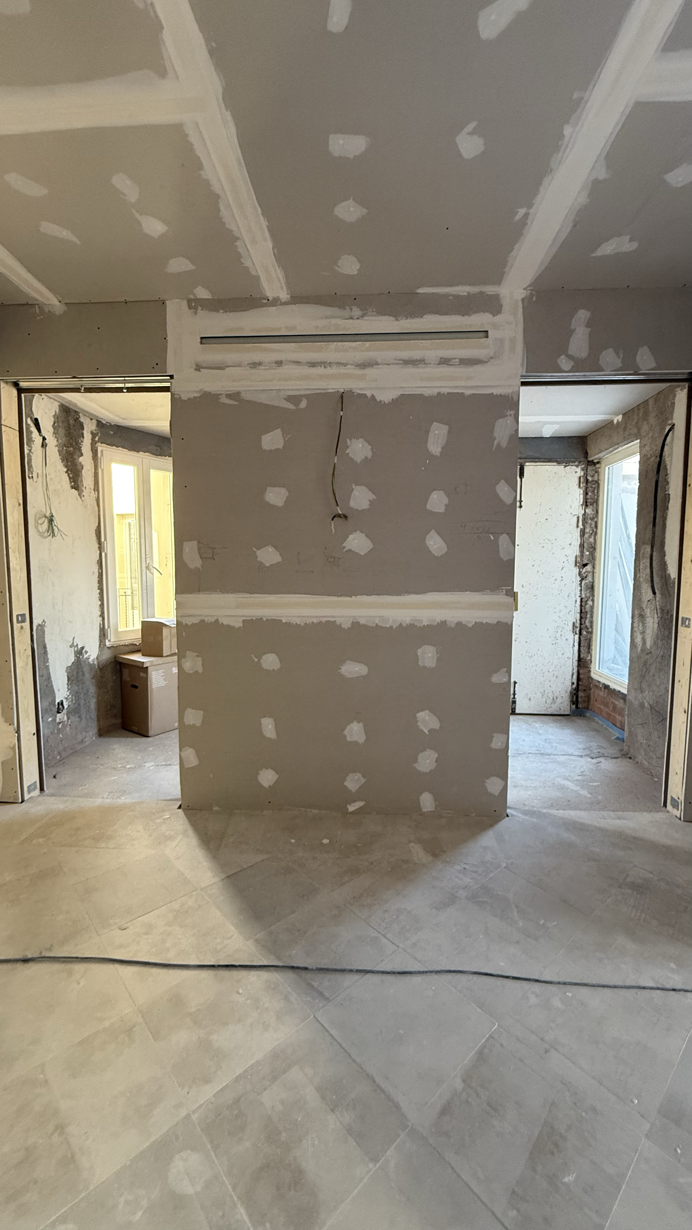 Interior of a house under construction with partially finished drywall and unpainted walls, with visible electrical wiring and large windows.