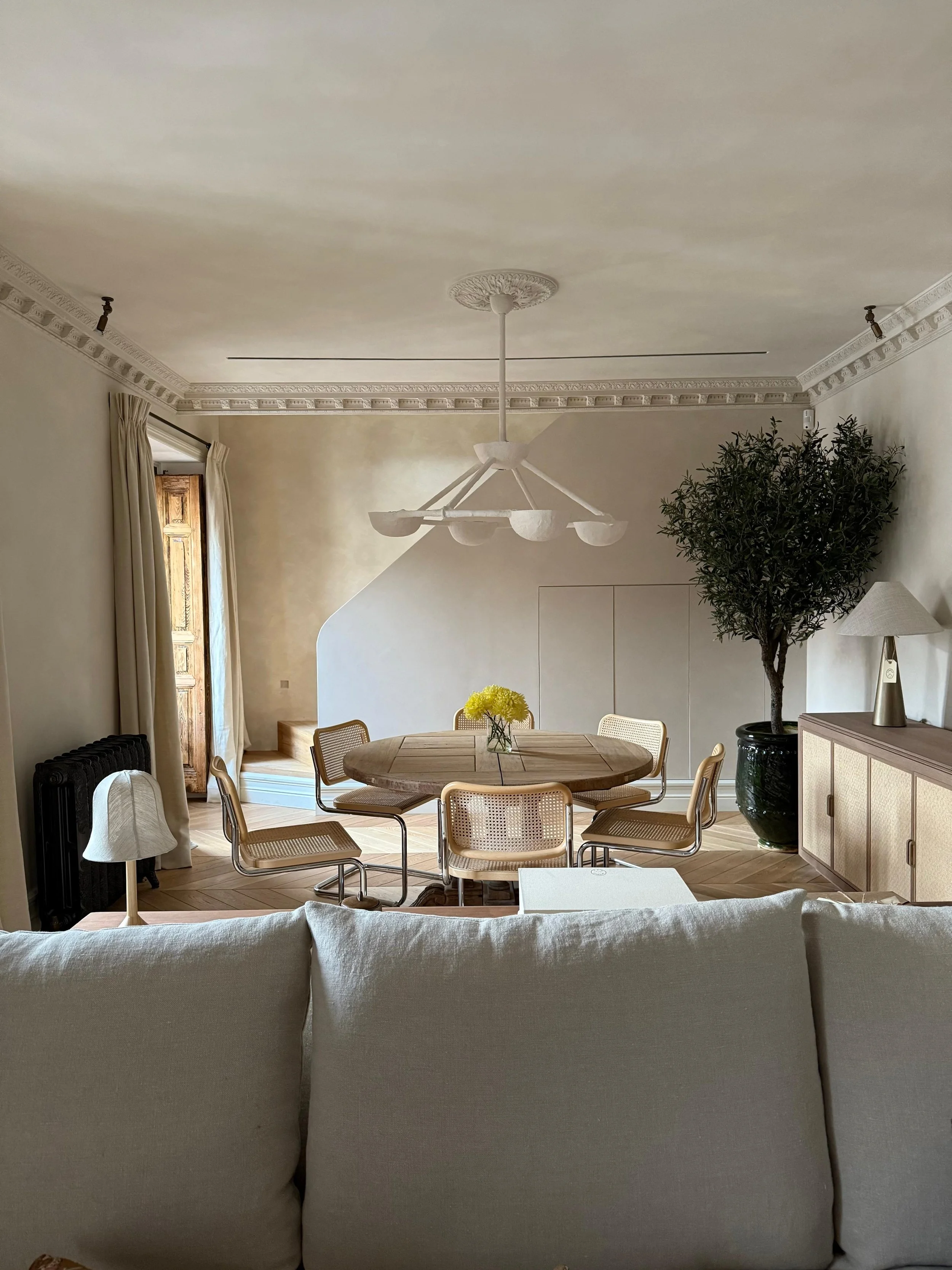 Living room with beige walls and ceiling, featuring crown molding, a circular wooden dining table with six chairs, a glass vase with yellow flowers, a large potted tree, a sideboard, a lamp, a ceiling light fixture, and beige curtains opening to a wooden door.