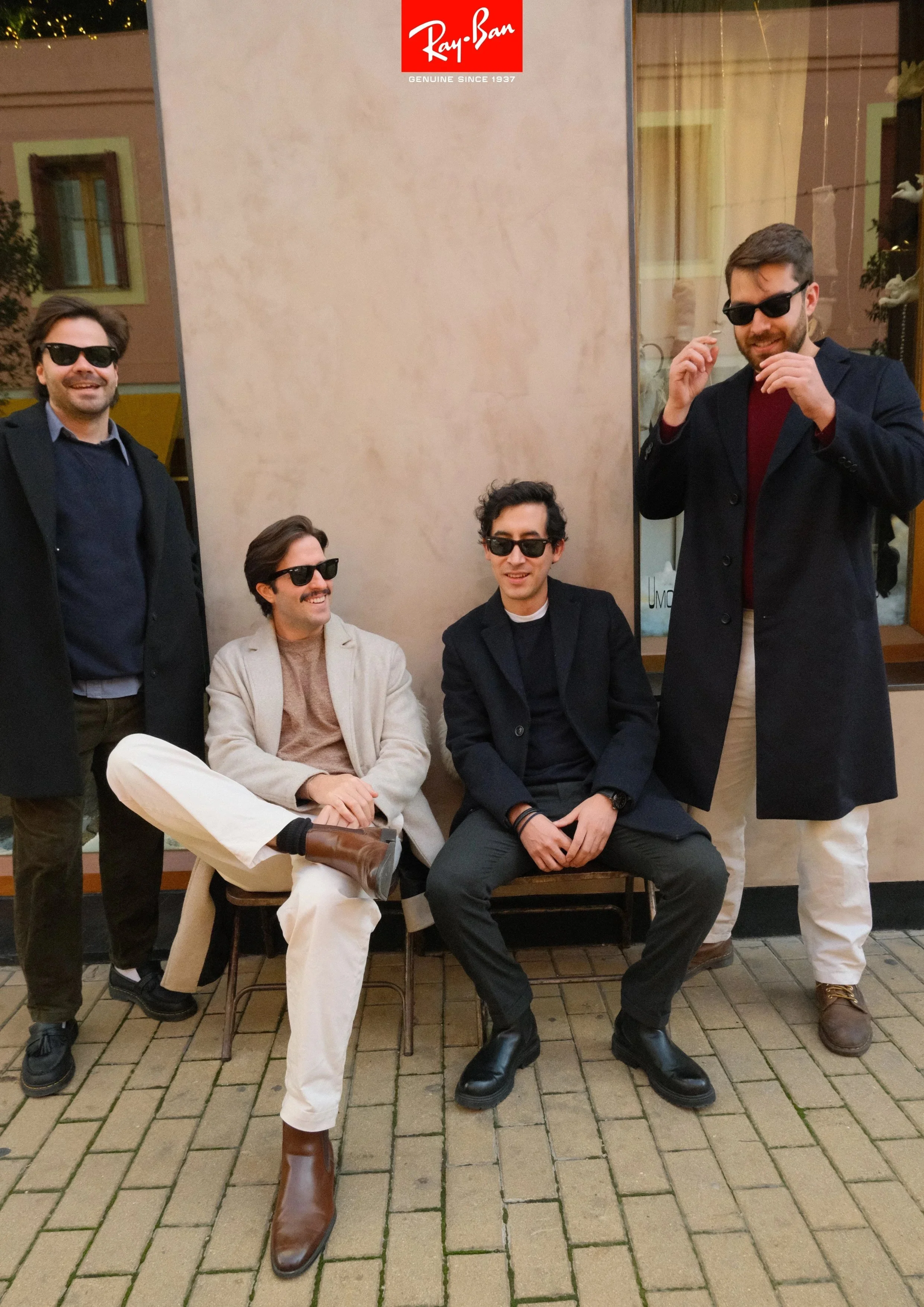 Four men with sunglasses posing in front of a Ray-Ban sign, some sitting and some standing outside on a brick sidewalk.