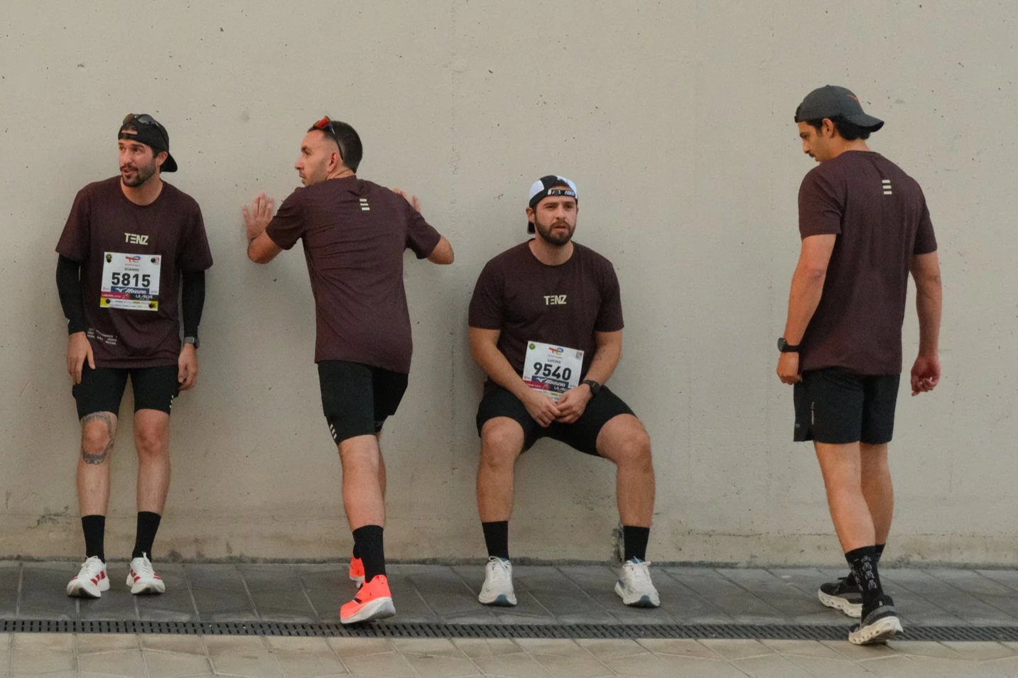Four men in matching maroon shirts and black shorts are taking a break against a plain wall. Two of them are standing and leaning against the wall, one is sitting on a chair, and the other is standing nearby. All are wearing running shoes and have race bibs on.