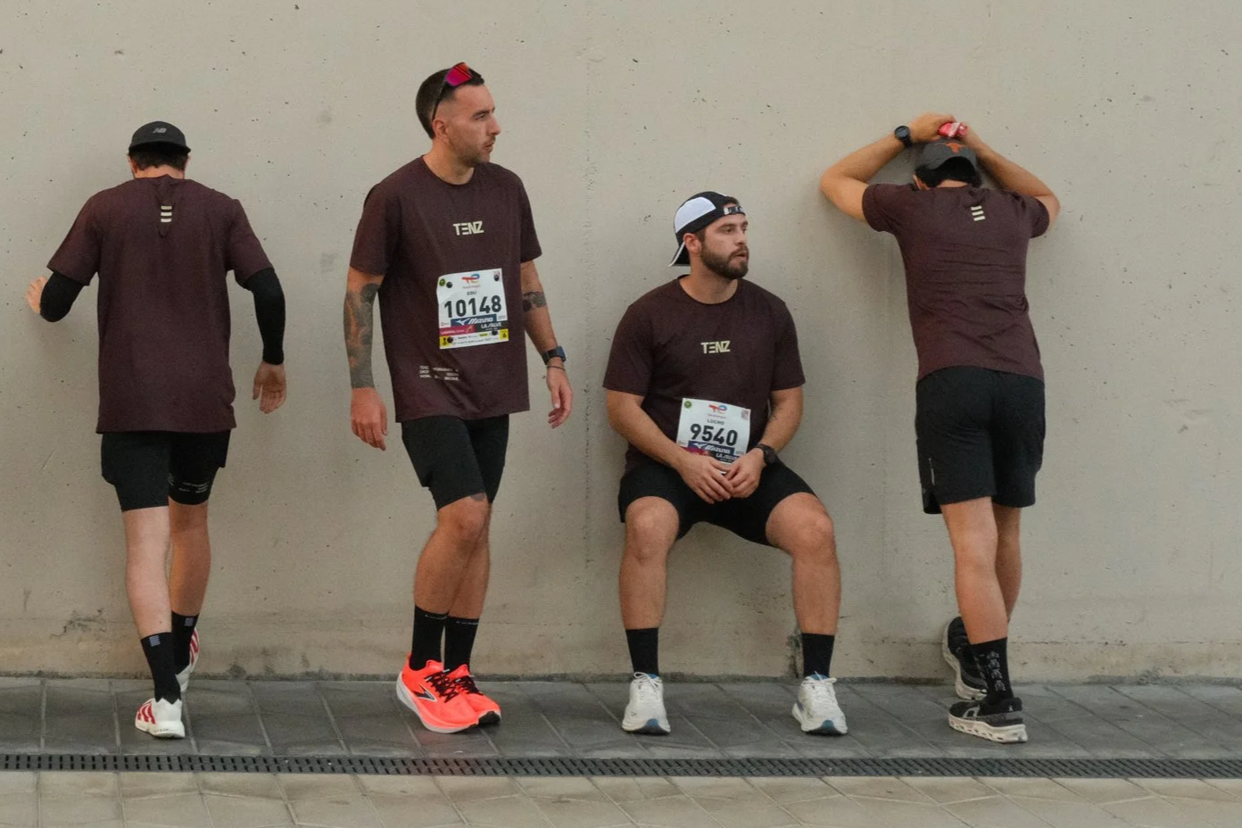 Four men in athletic wear are waiting against a plain wall, with one sitting and the others standing, some with their backs turned or resting their heads.