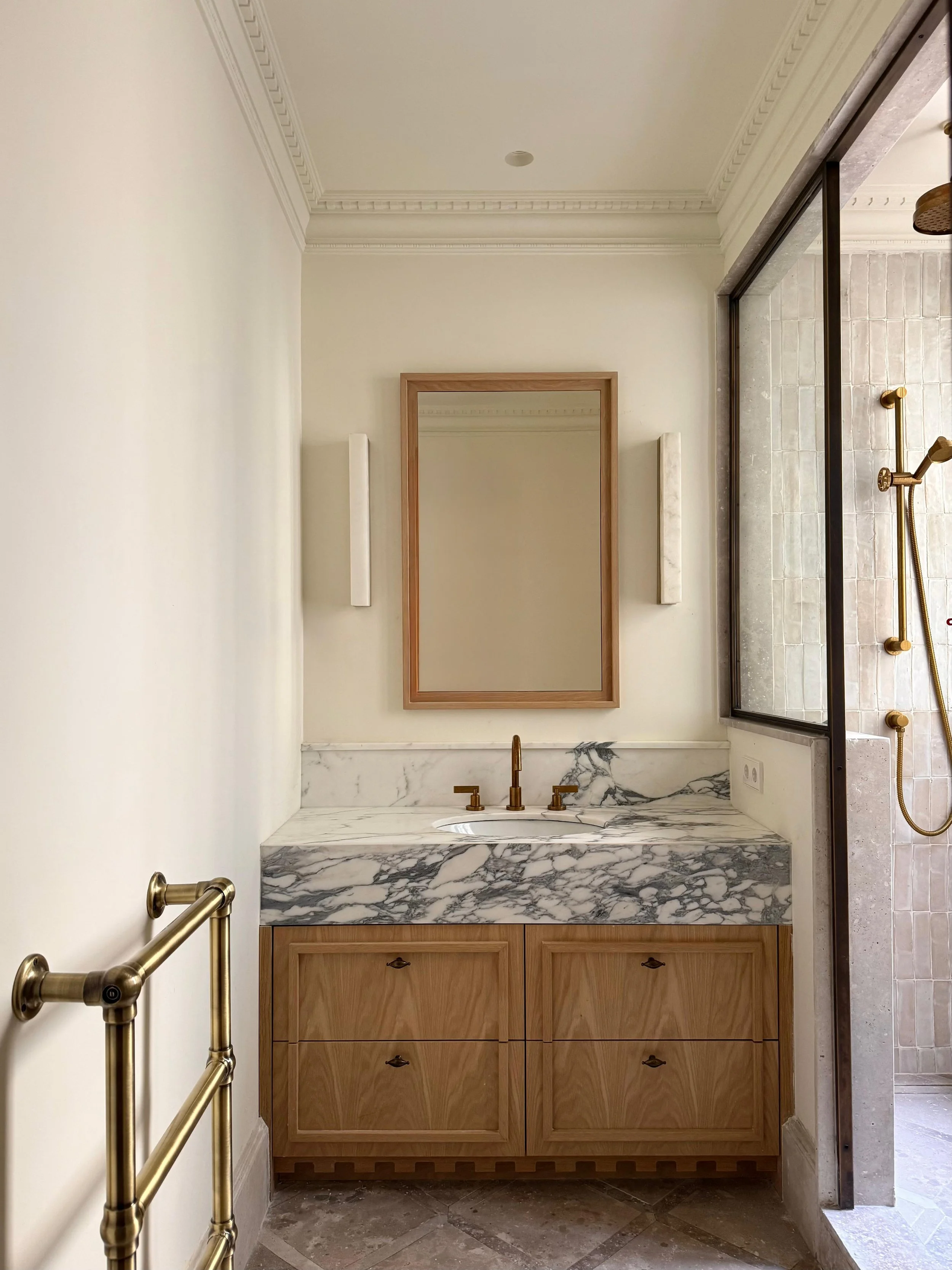 Bathroom vanity with a marble top, wood cabinets, a mirror, and a brass faucet. There is a glass shower to the right with chrome fixtures and a wall-mounted towel rack to the left.