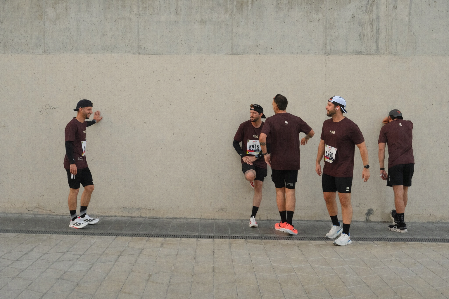 Five men in athletic clothing and race bibs talking and smiling near a plain beige wall with graffiti that says 'I ♥ data'