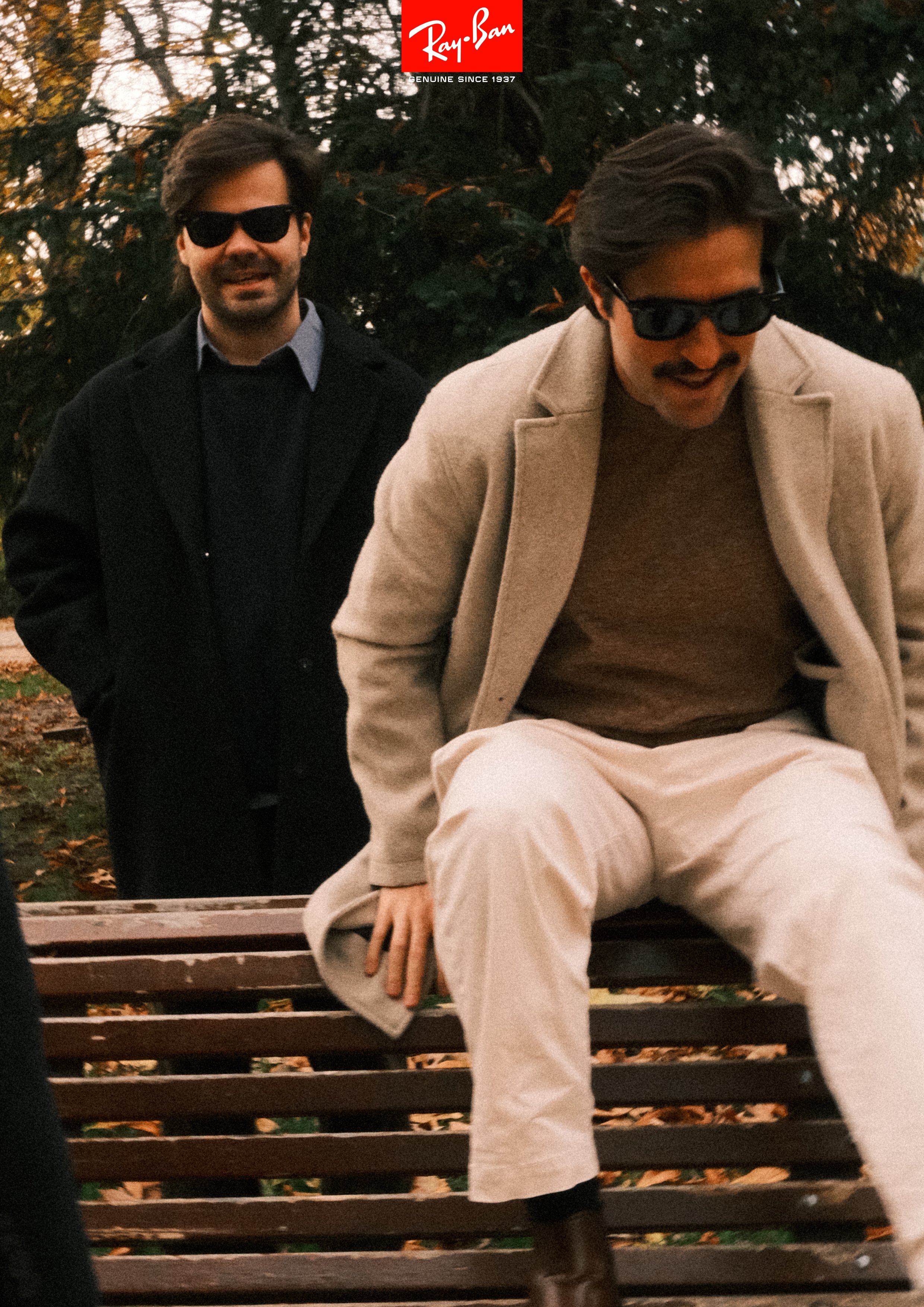 Two men with sunglasses in a park with autumn leaves. One man sits on a bench while the other stands behind him.