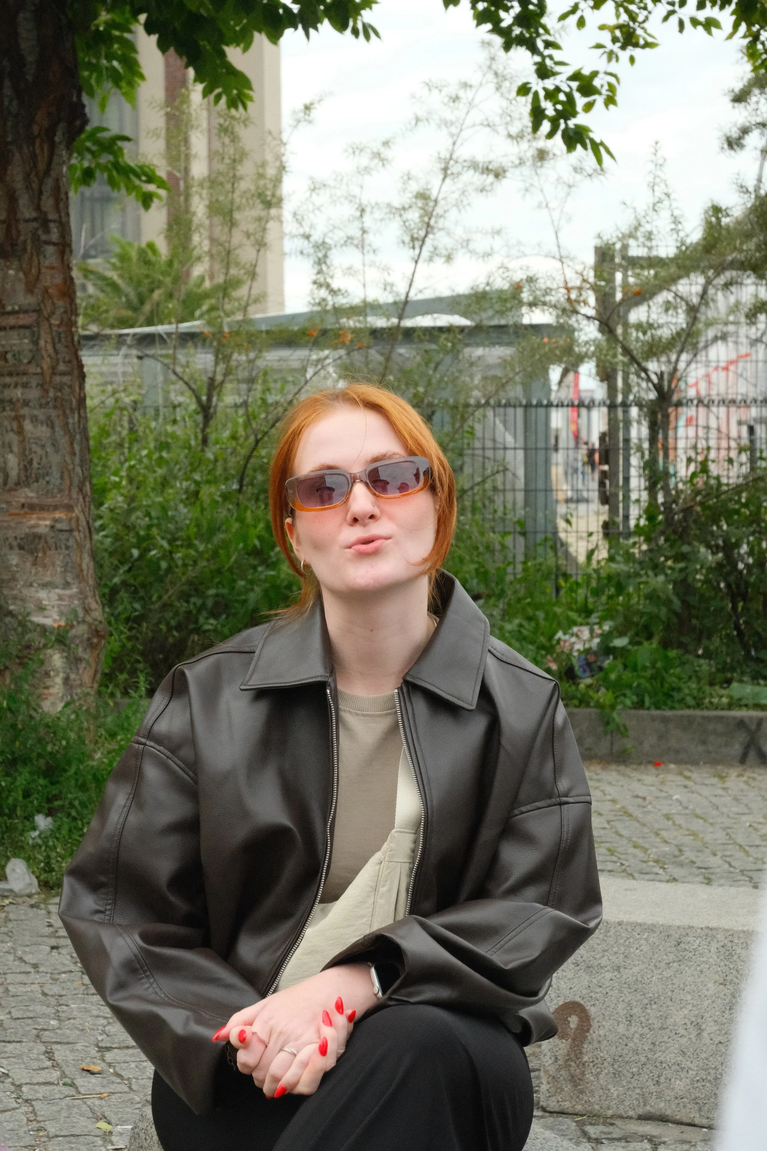 A woman with short red hair wearing sunglasses, a dark leather jacket, a beige shirt, and red nail polish, sitting outdoors on a concrete bench with greenery and trees in the background.