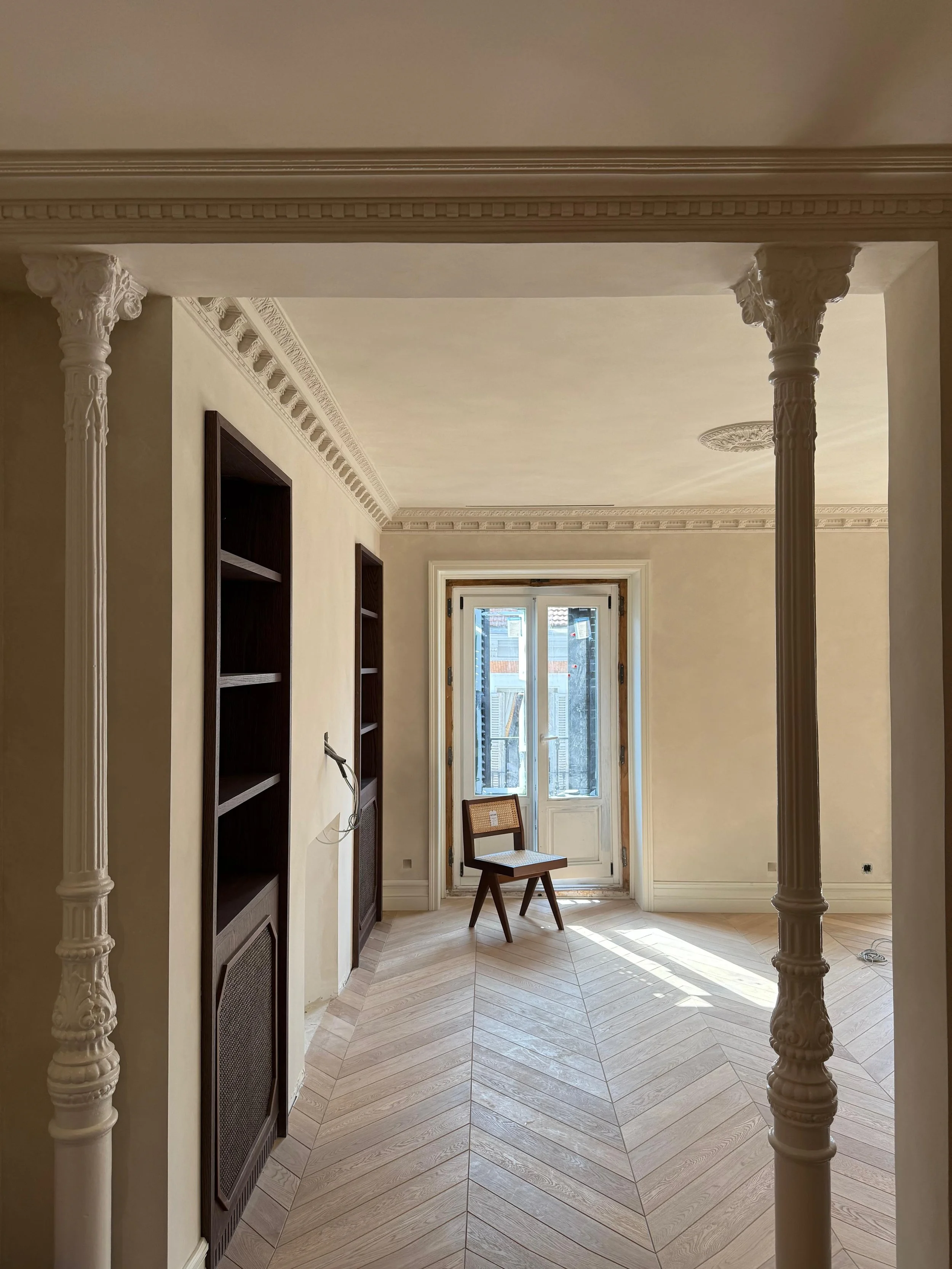 An empty room with ornate architectural details, including decorative columns and crown molding. There is a window or door at the far end letting in natural light, with a wooden chair placed in front of it. Built-in dark wood bookshelves are on the left side of the room, and the floor has a herringbone wood pattern.
