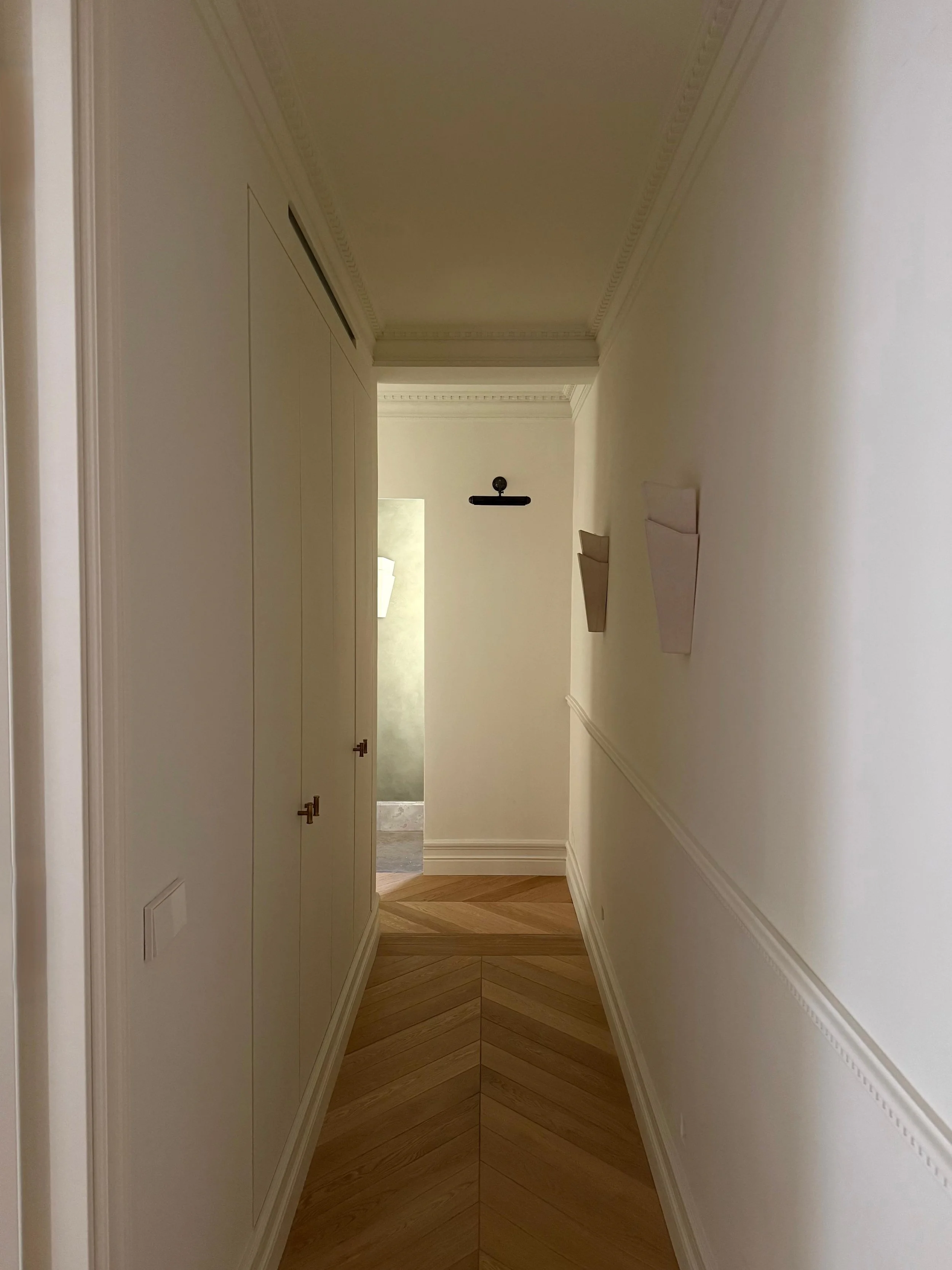 A narrow hallway with white walls, wooden chevron-patterned flooring, and built-in white cabinets on the left side. There is a wall-mounted shelf and decorative wall molding. At the end, an open doorway reveals a glimpse of another room with a wall-m