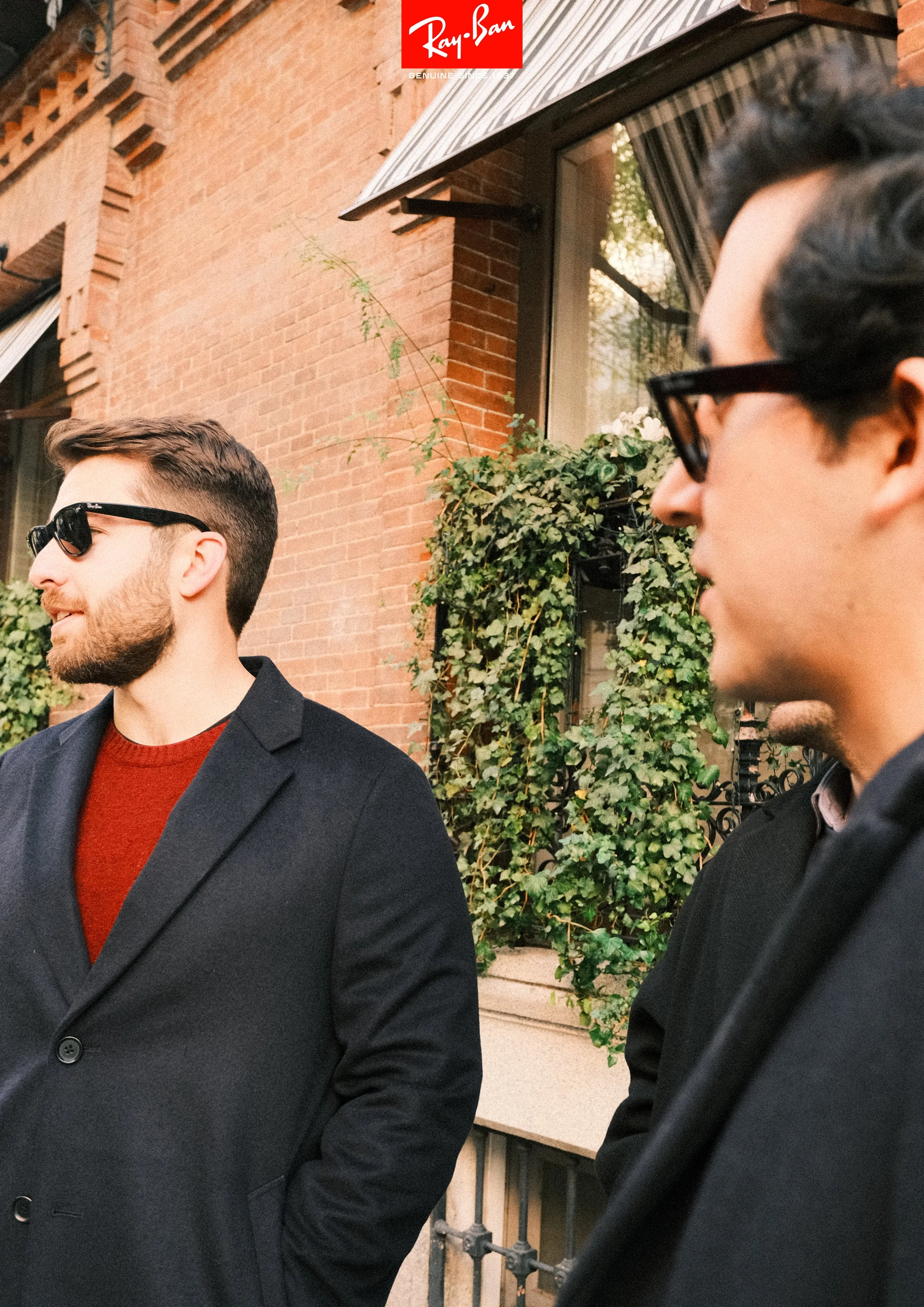 Two men standing outside in front of a brick building with greenery, both wearing black coats and sunglasses, engaged in conversation.