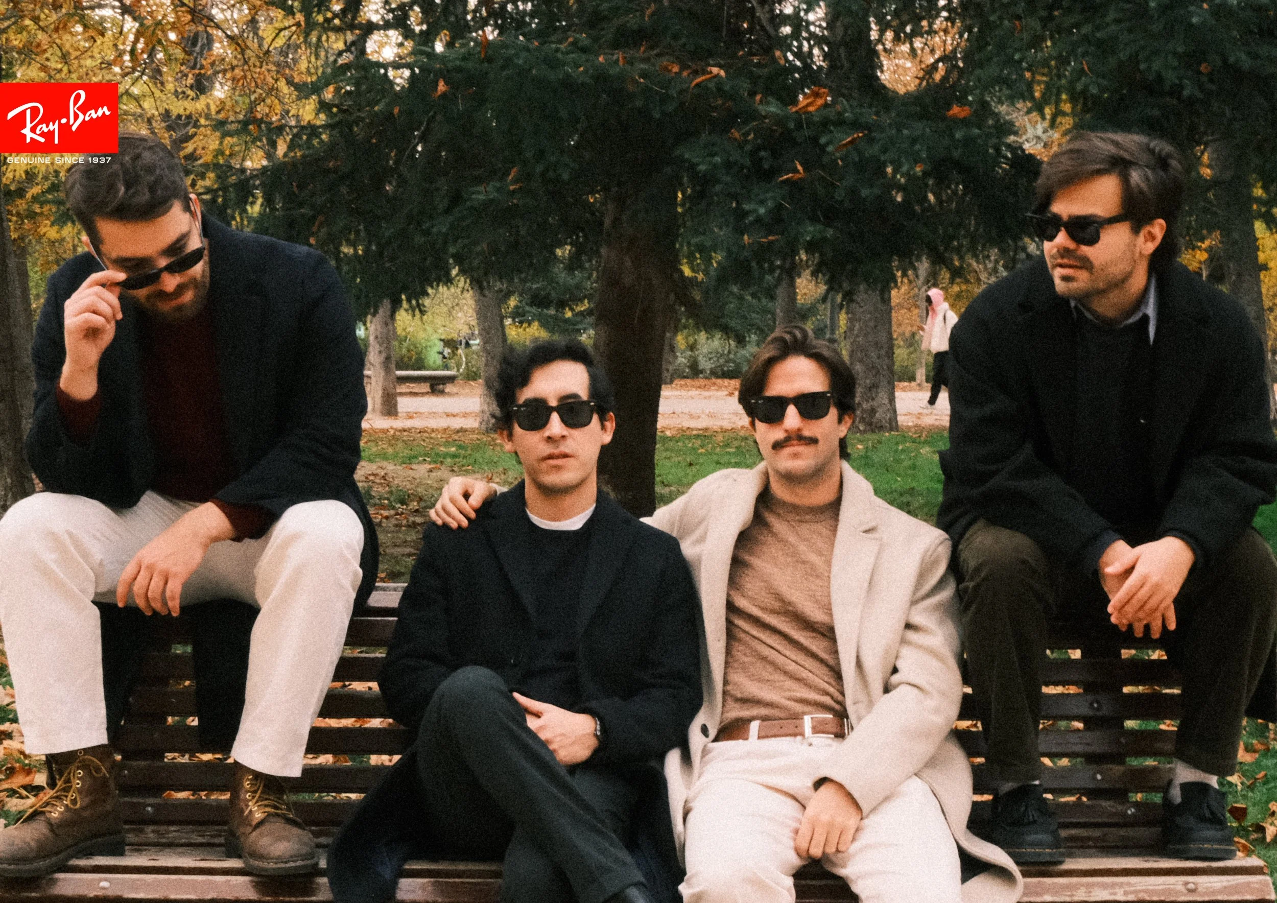 Four men sitting on a park bench wearing sunglasses, with trees and fallen leaves in the background. The logo for Ray-Ban is in the top left corner.