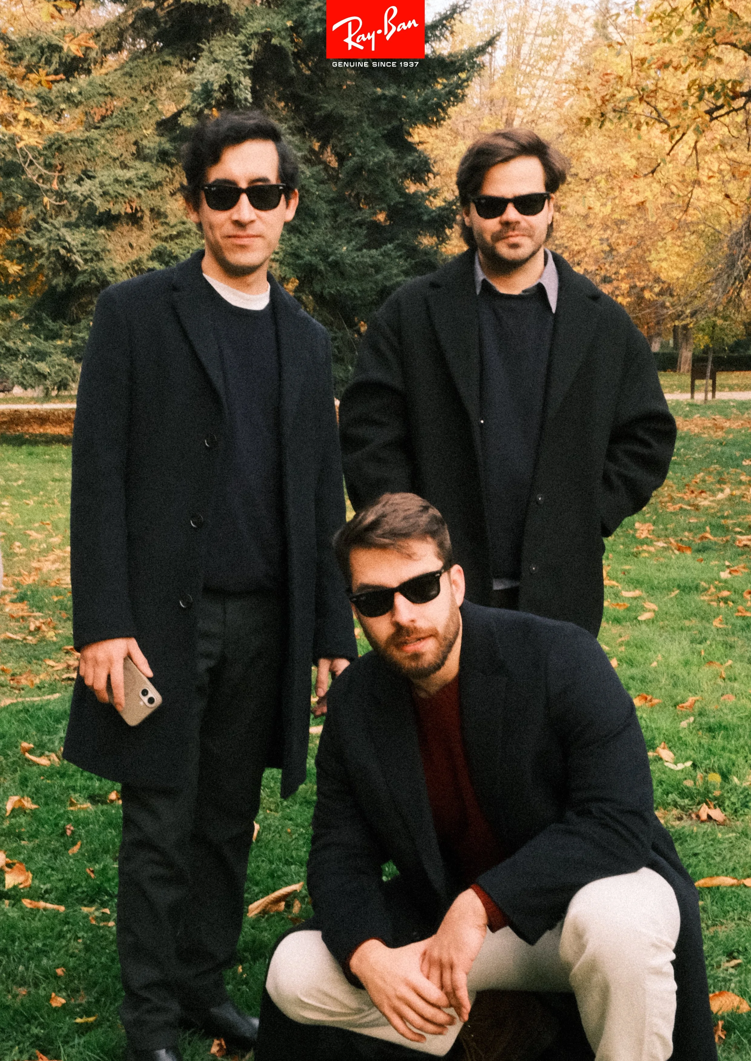 Three men wearing dark sunglasses and black coats, posing outdoors in a park with autumn leaves and trees in the background.