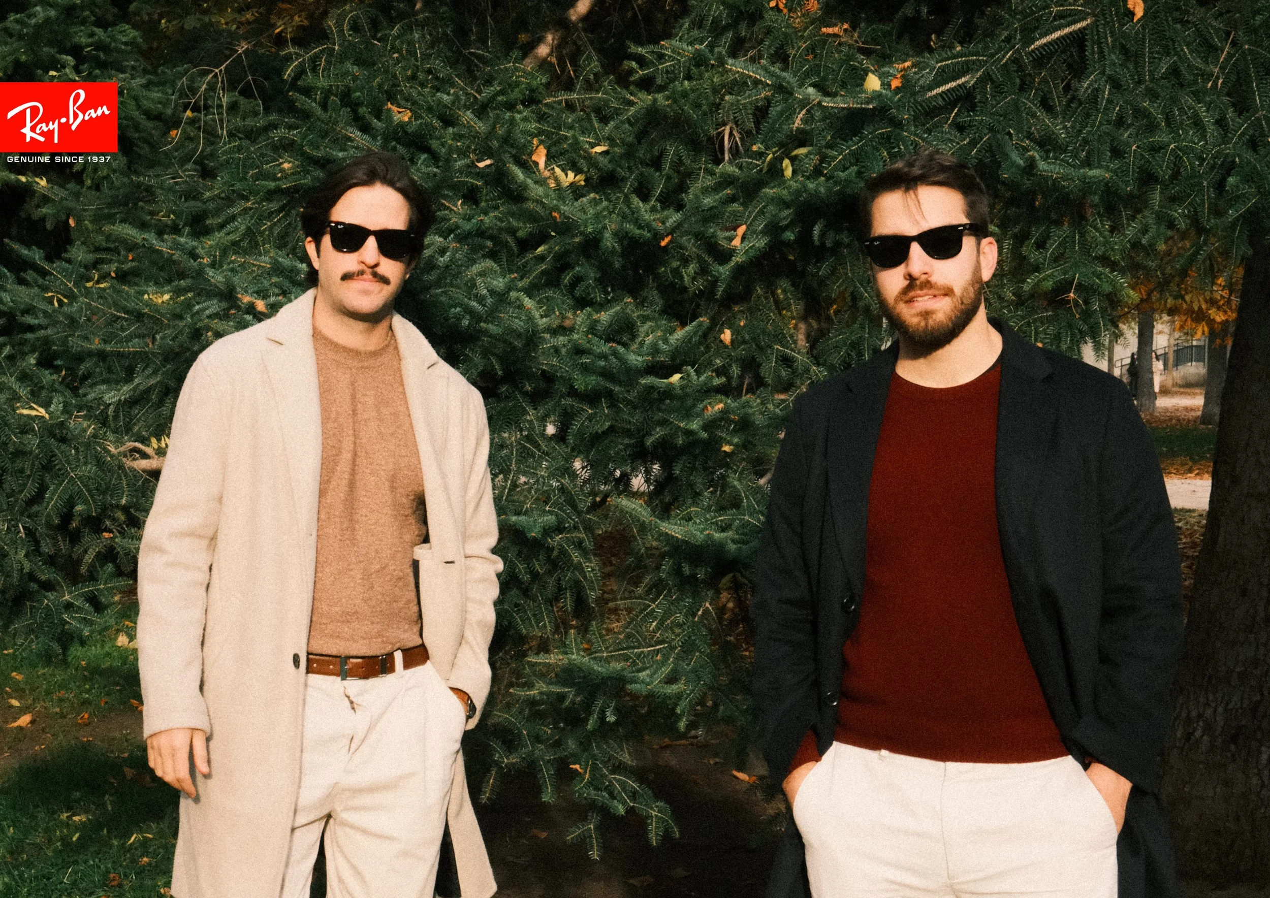 Two men standing outdoors near a large Christmas tree. Both are wearing sunglasses, with the man on the left dressed in a beige coat, brown sweater, and light-colored pants, and the man on the right in a black blazer, red shirt, and white pants.