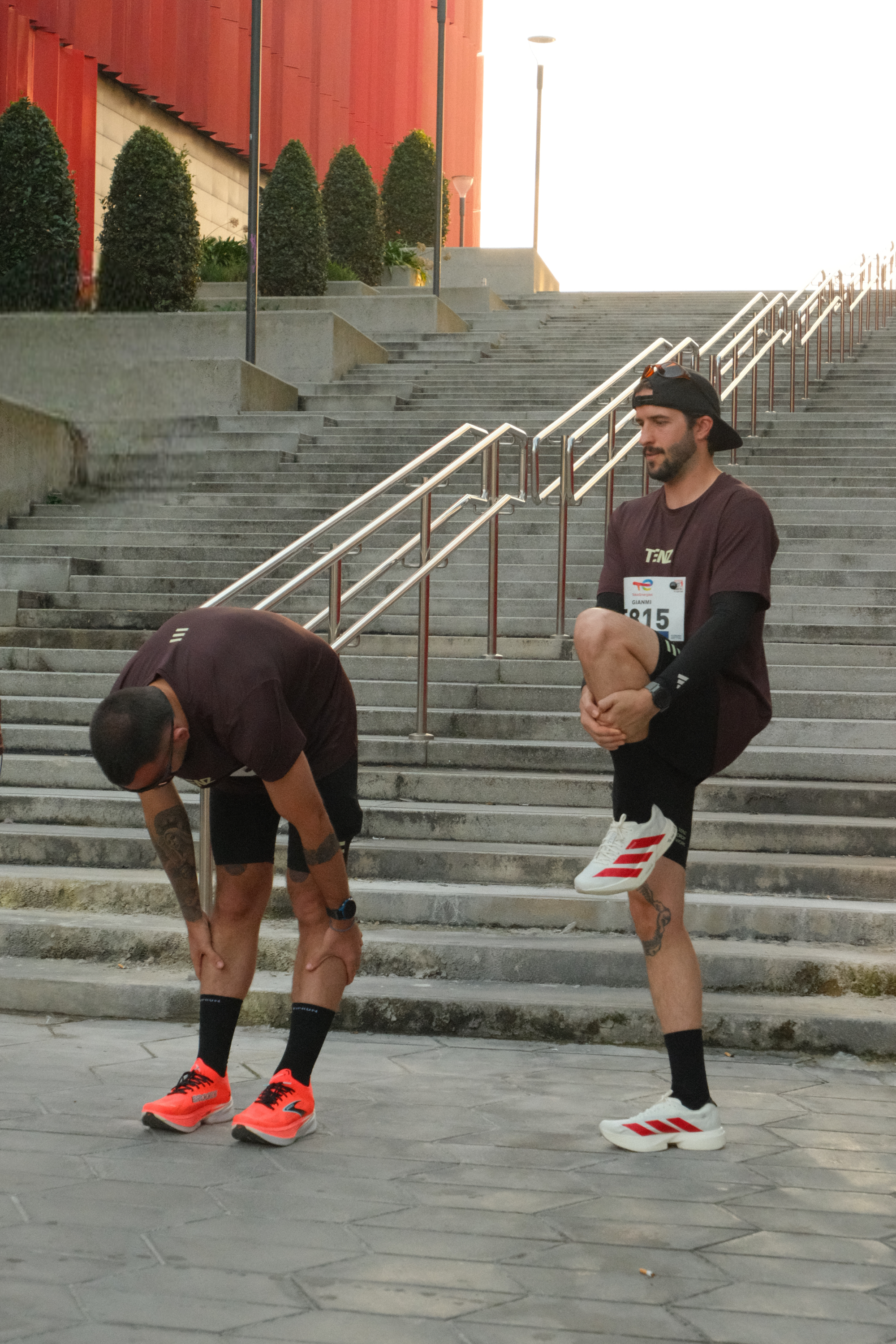 Two men in athletic clothing stretching on city stairs. One man is bending forward, touching his toes, while the other man is standing on one leg and holding his ankle behind him.