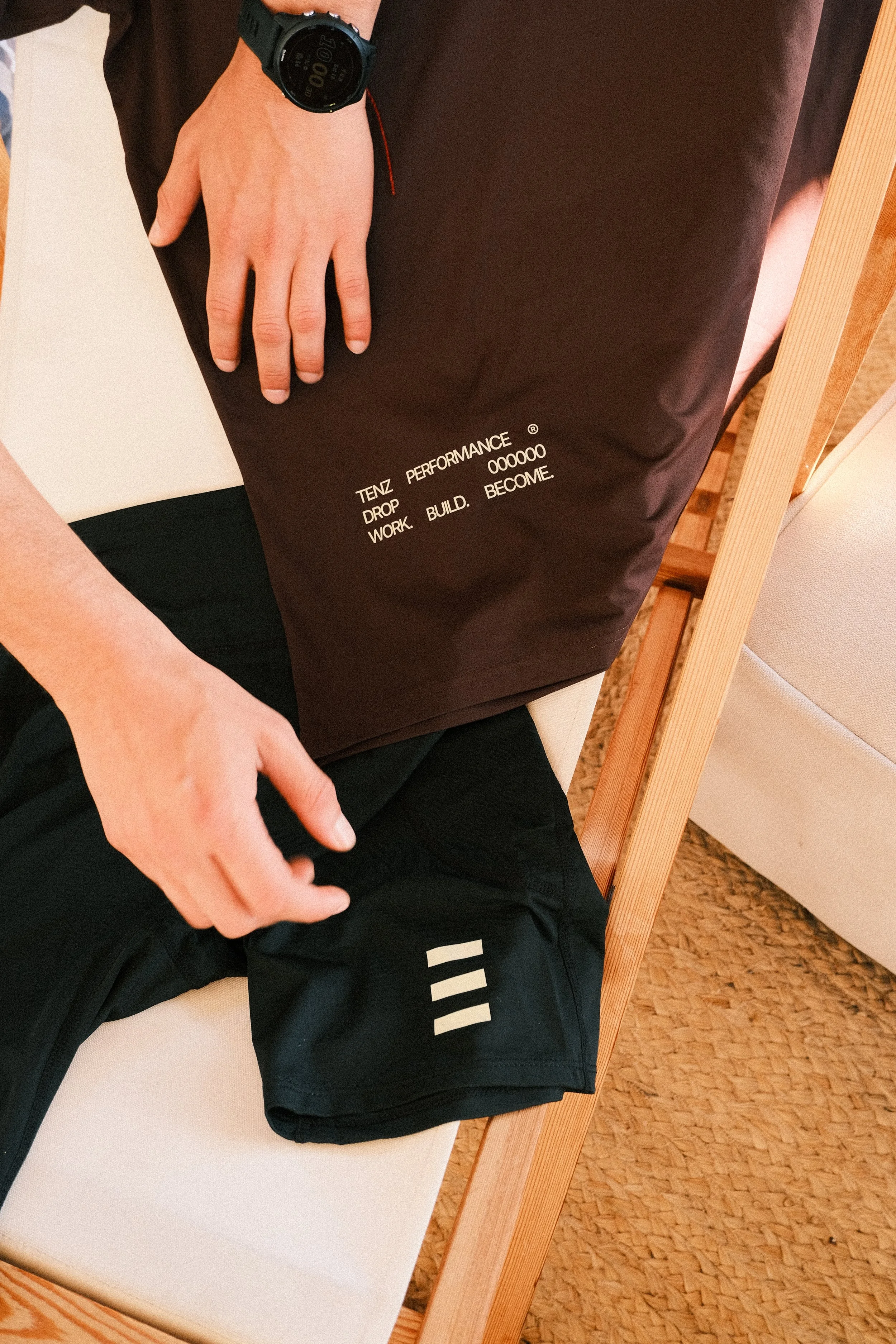 Person sitting on a bench holding a dark brown bag with white text, wearing a black watch and black athletic shorts with white stripes, with part of a white chair and beige carpet visible.