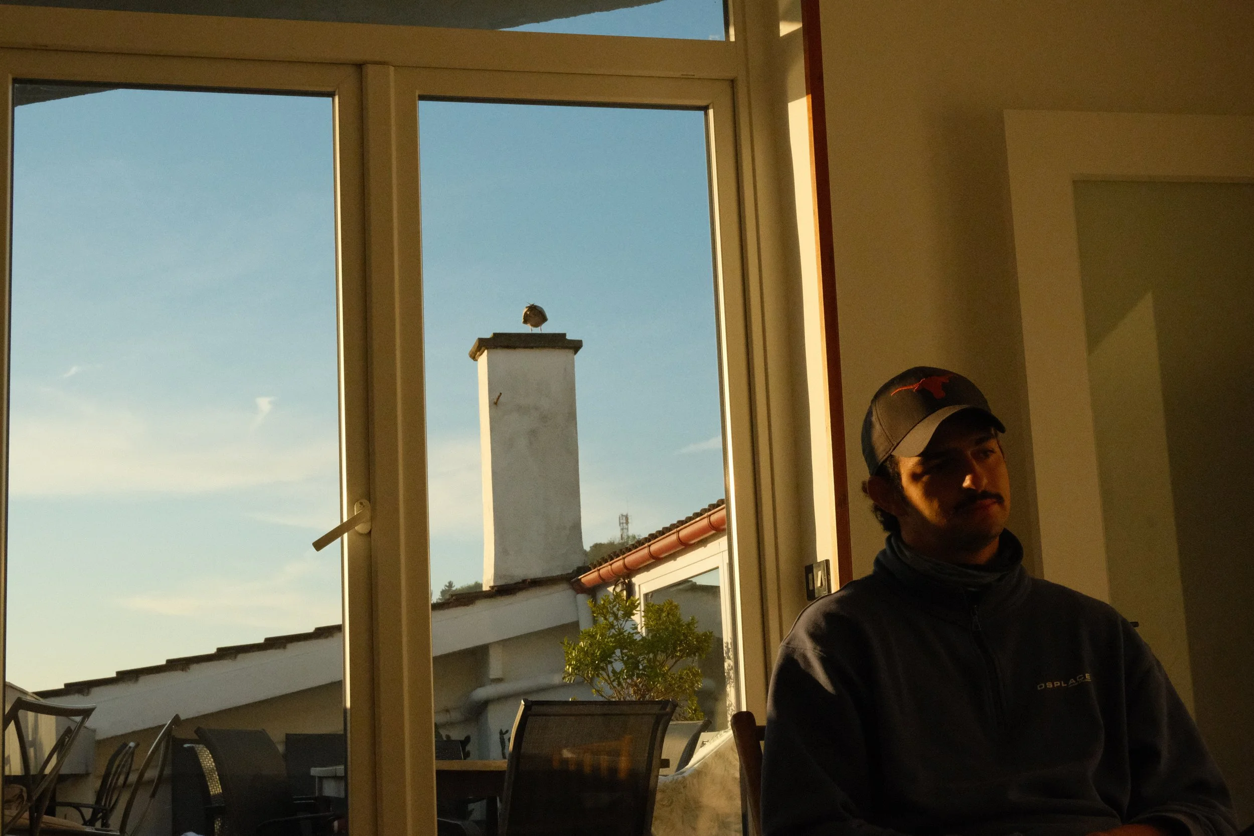 Man sitting indoors near a window with outdoor view of chimney and roof at sunset.