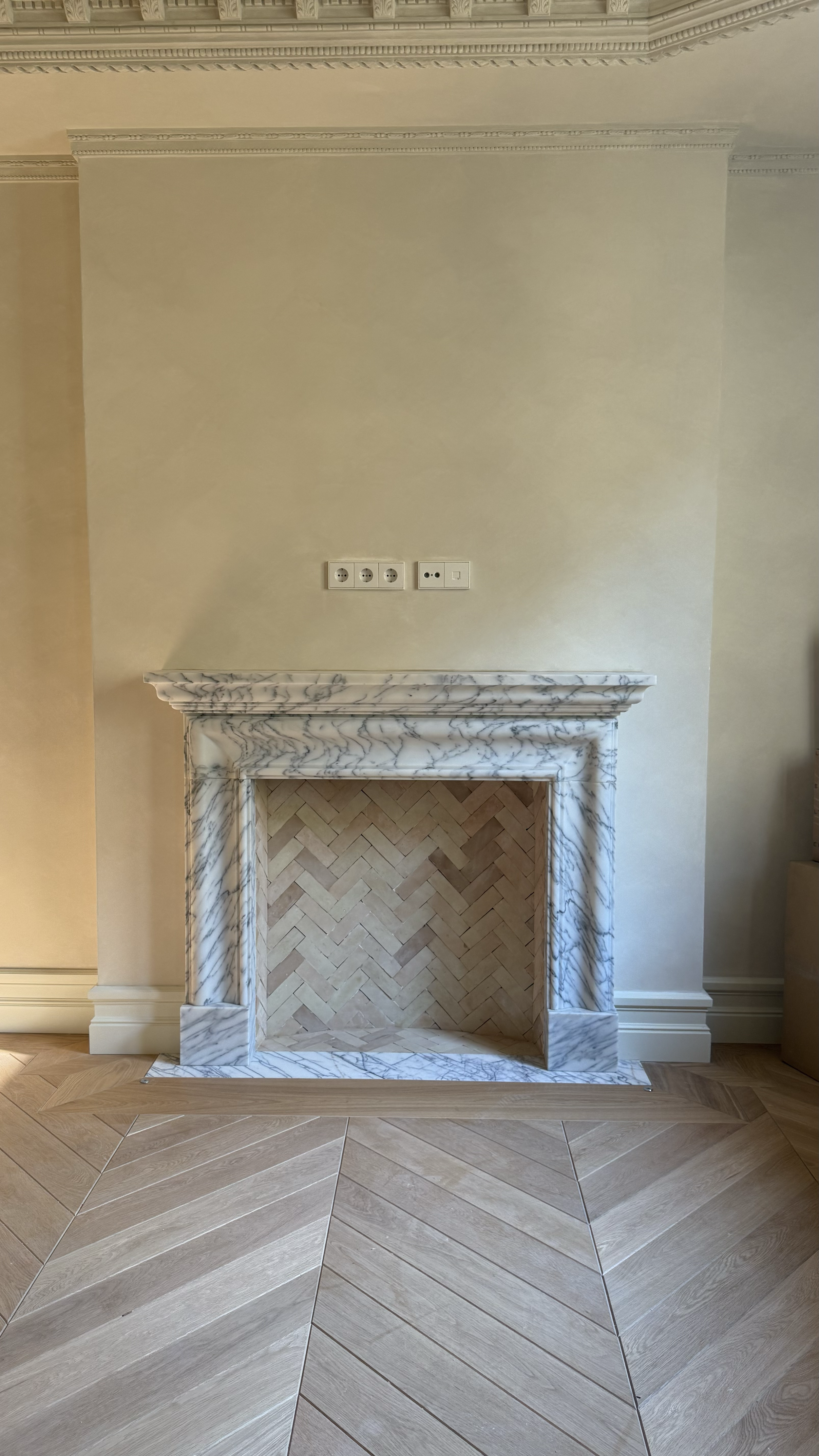 A decorative fireplace with a marble frame, a plain beige wall with electrical outlets above, and parquet flooring in front.