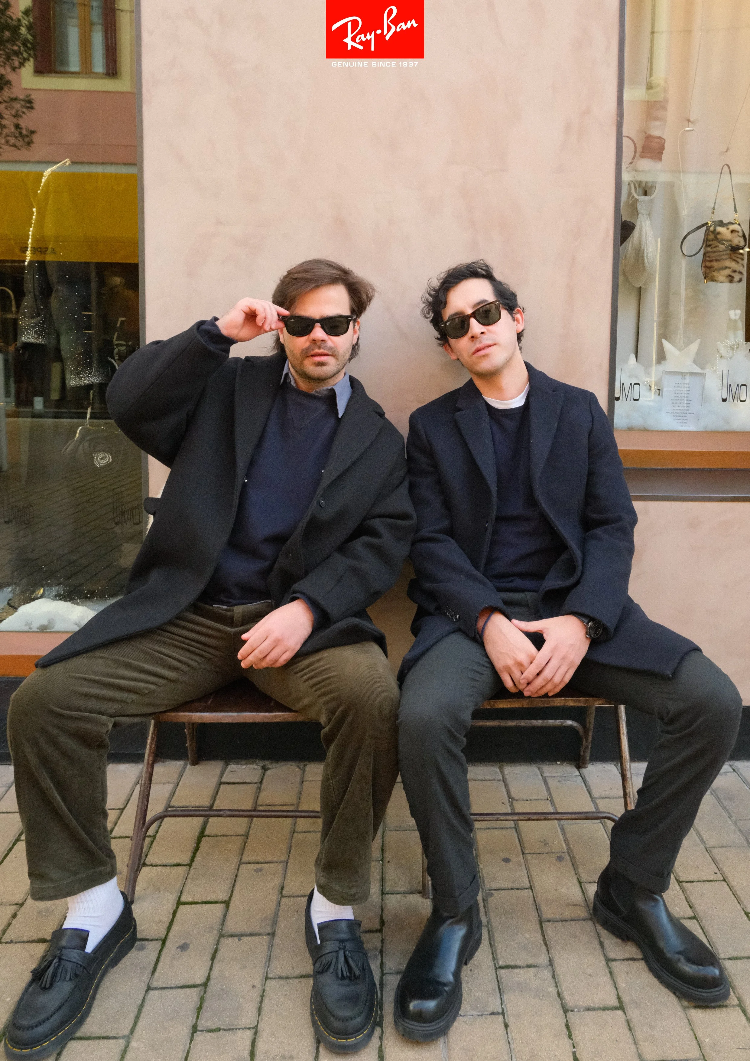 Two young men sitting on a bench outside a store with a Ray-Ban sign, both wearing dark sunglasses and stylish black jackets. One adjusts his sunglasses while the other leans back with hands on his lap. The background shows a window display with vari
