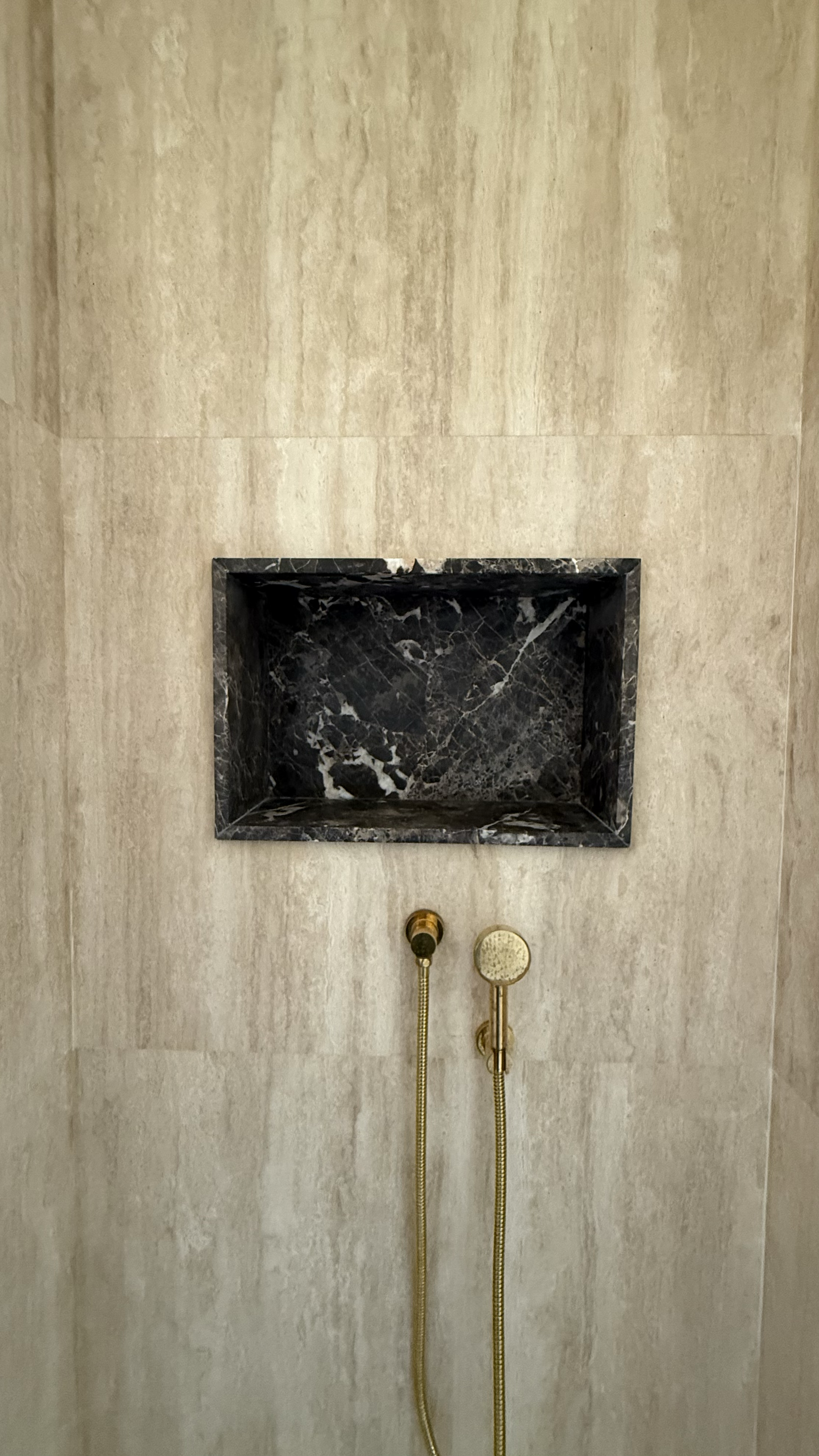 Shower with black marble shelf, gold handheld showerhead, and beige tiled walls.