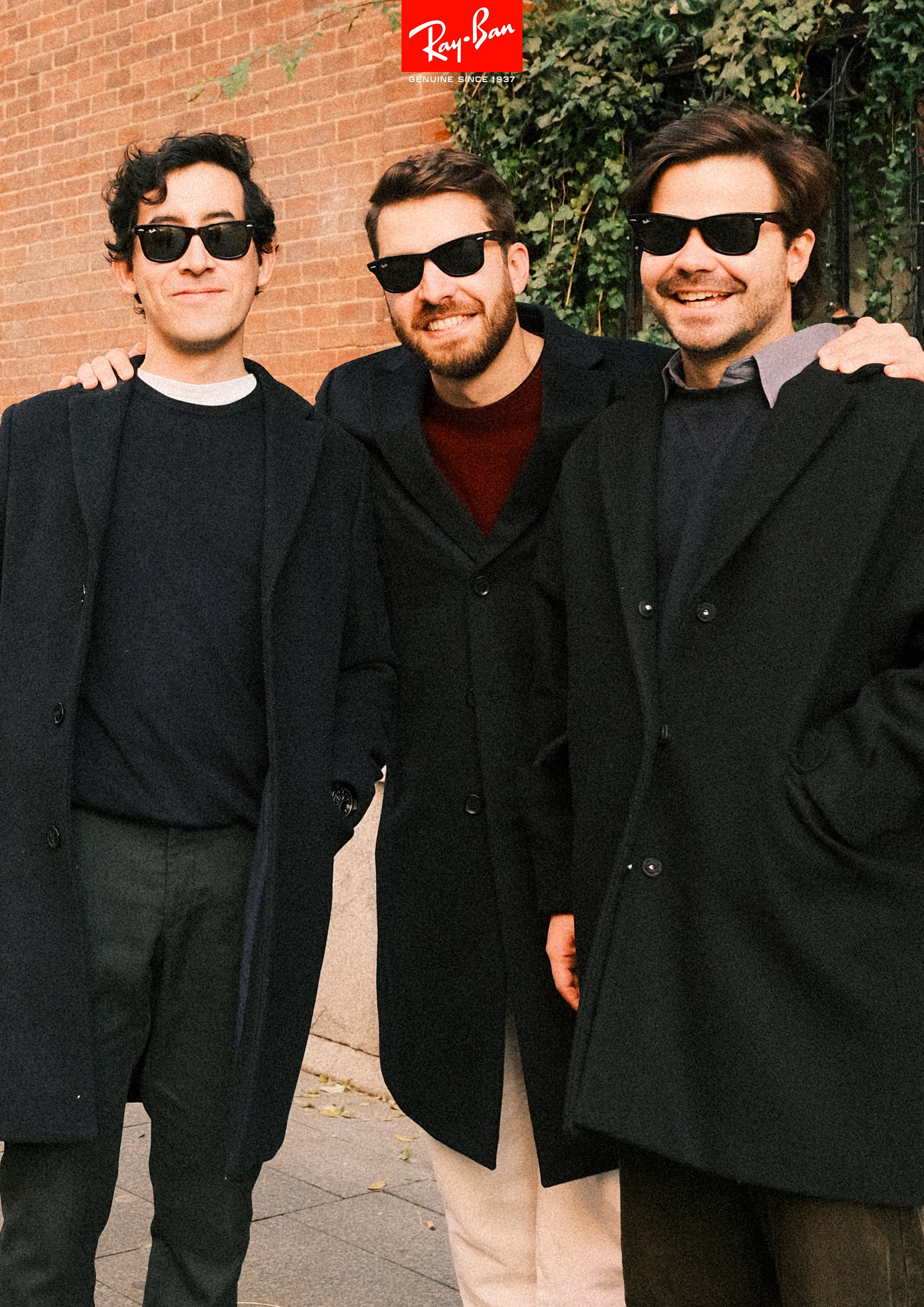 Three men standing outdoors in front of a brick wall and green ivy, all wearing black coats and sunglasses, smiling at the camera.