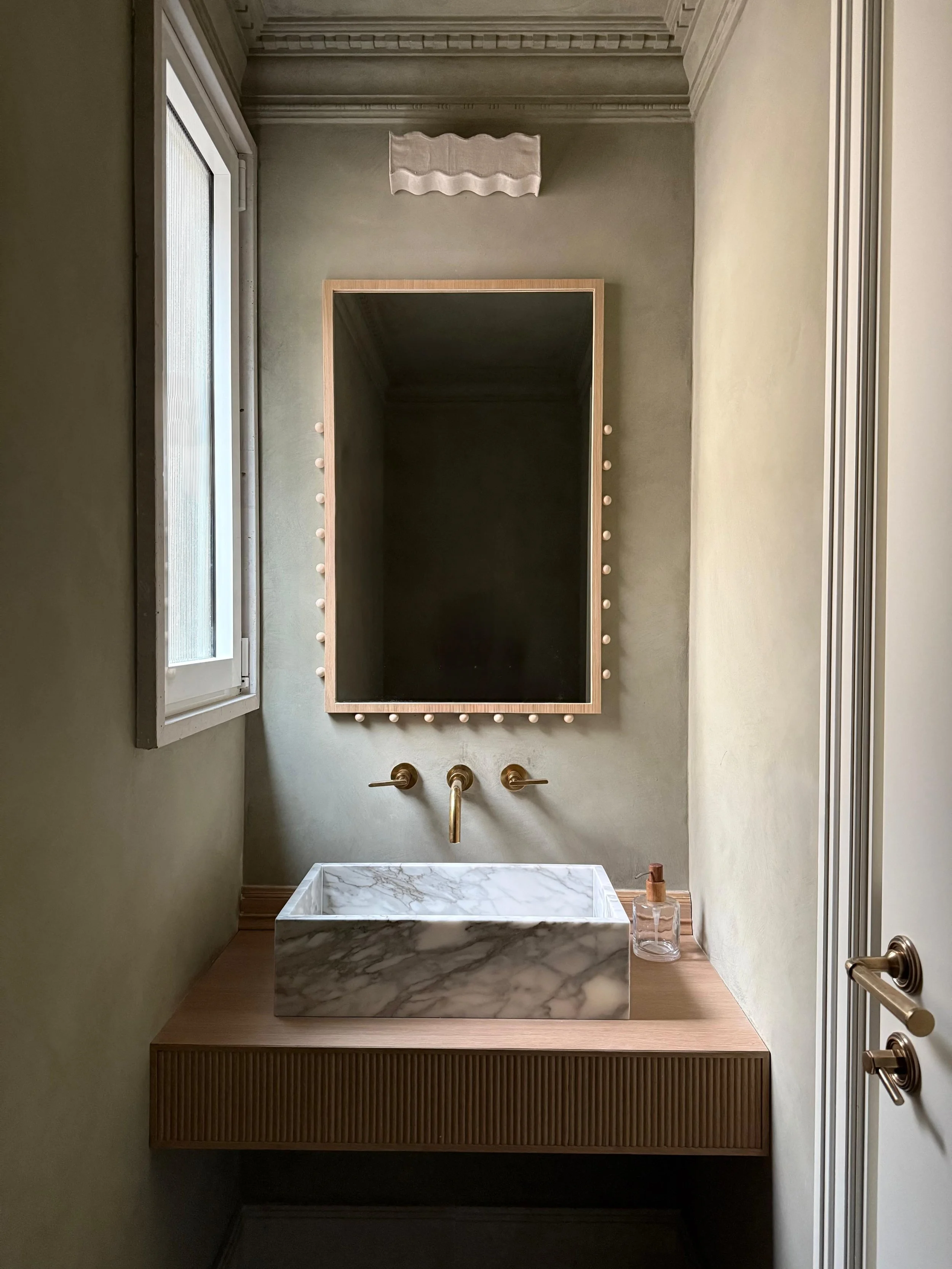 Small bathroom with a marble sink, a wooden vanity, a mirror with a wooden frame and light bulbs, a wall-mounted faucet, a window on the left, and a bottle of liquid soap on the right.