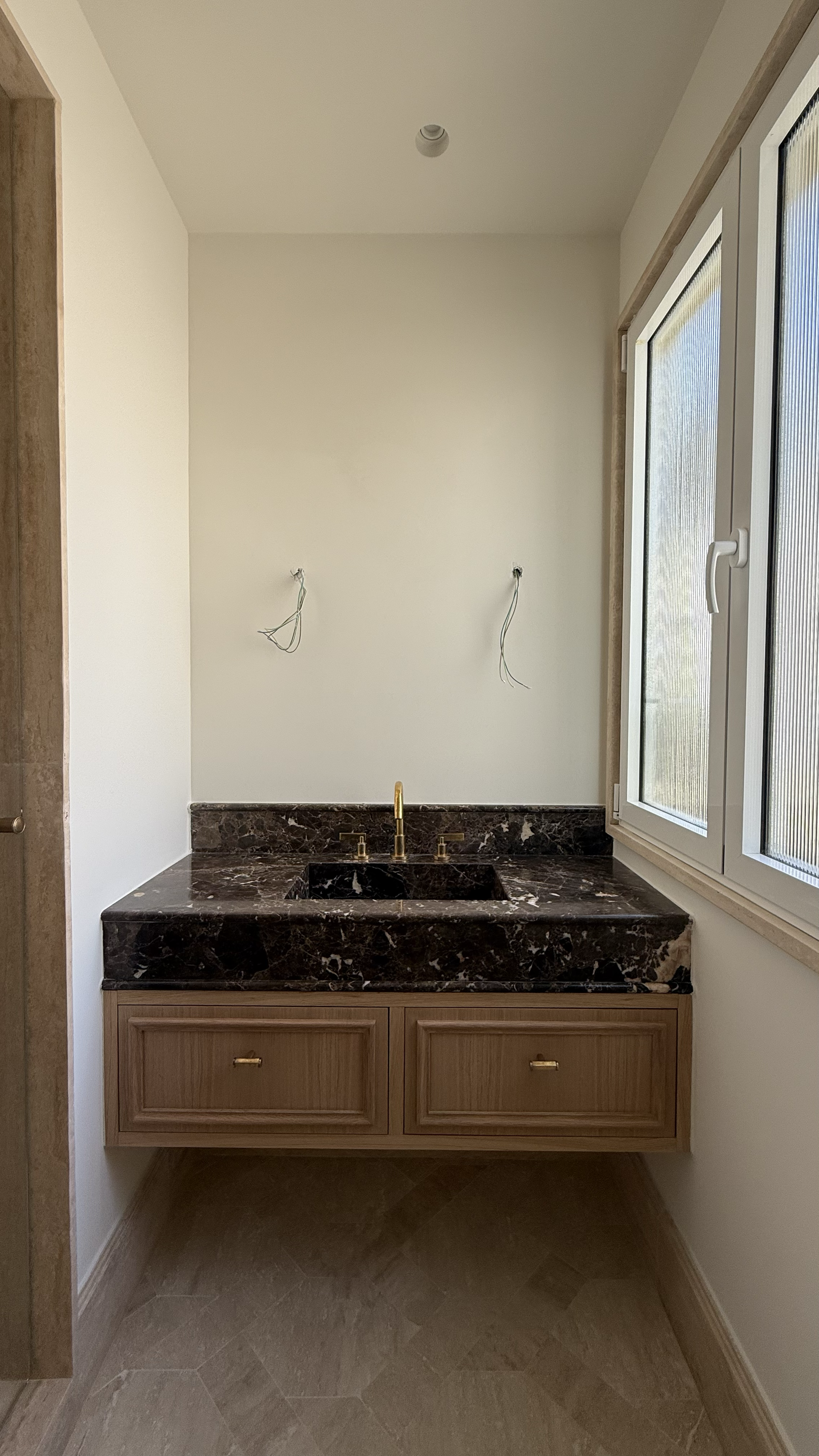 Interior of a bathroom under construction with a marble countertop, wooden cabinetry, and an uninstalled sink and fixtures, with wiring visible on the wall.