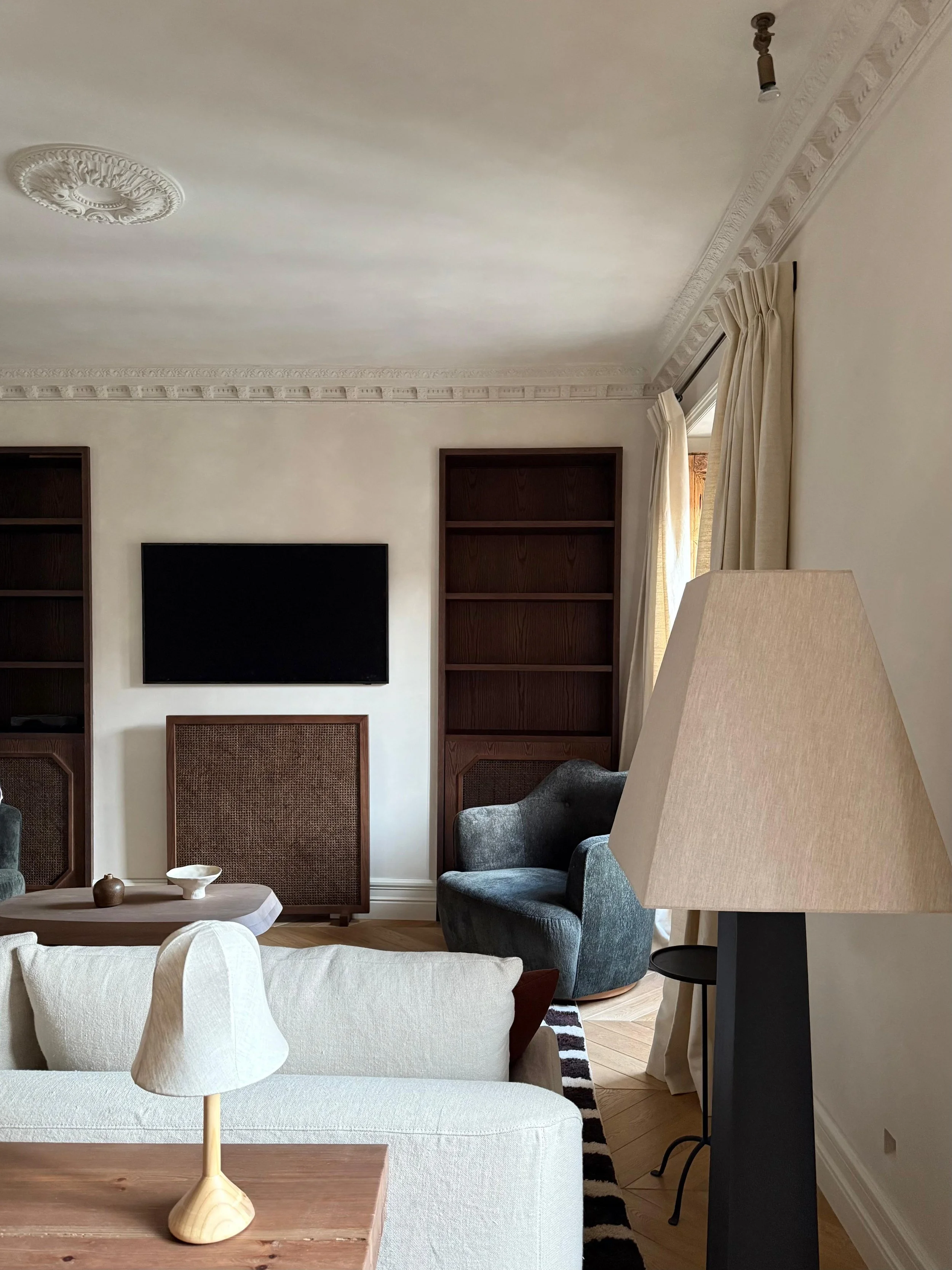 A cozy living room with a white sofa, a small wooden side table with a beige table lamp, a gray armchair, dark wooden bookshelves, a wall-mounted flat-screen TV, a large window with cream-colored curtains, and a ceiling with ornate molding.