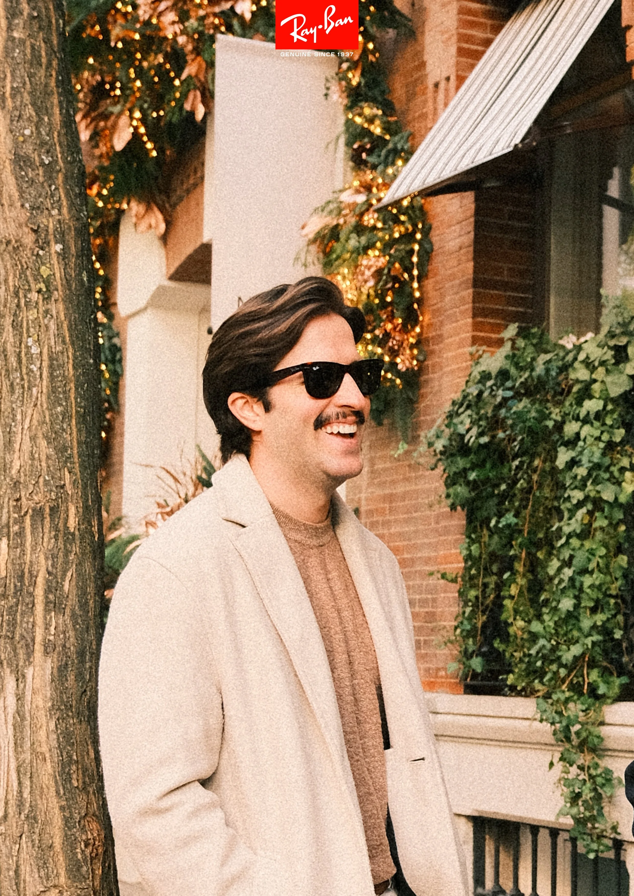 A man with a mustache and dark hair wearing sunglasses, a beige blazer, and a brown sweater, smiling outdoors near a tree, with Christmas lights and decorations in the background.