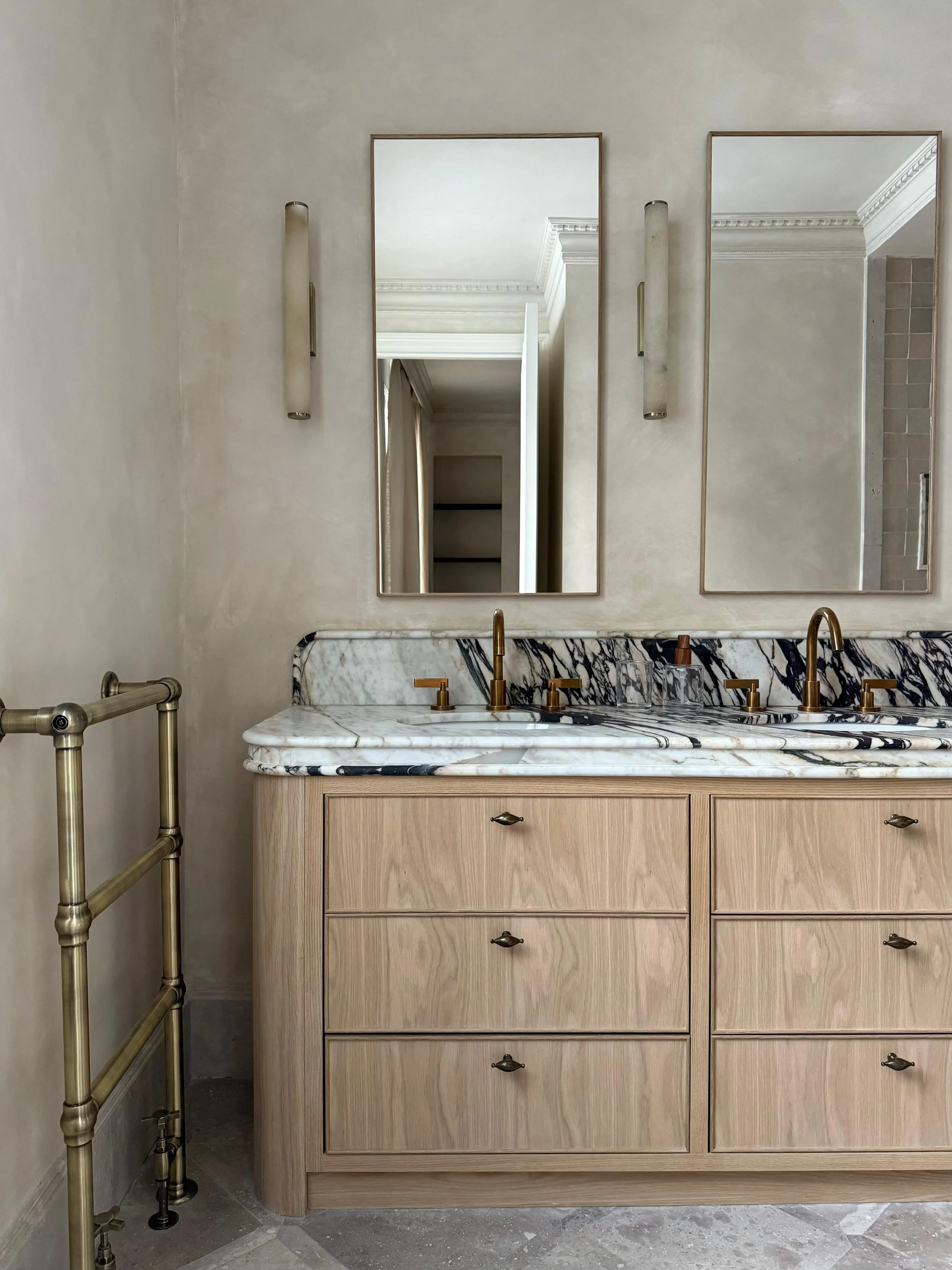 A bathroom vanity with a marble countertop, two mirrors, and two wall-mounted light fixtures, with a metal rail on the left side and a tiled wall in the background.
