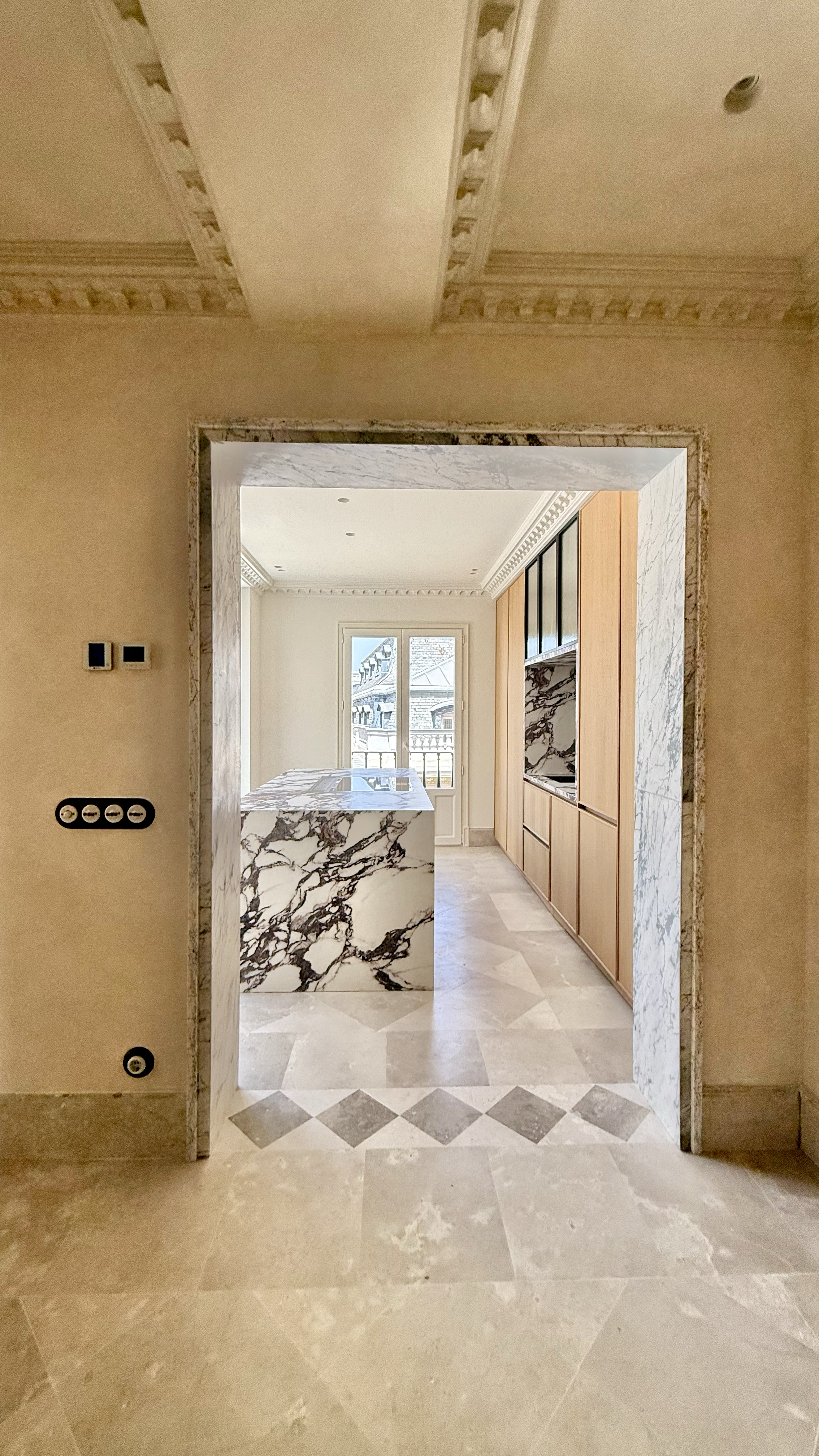 A view through a doorway into a modern kitchen with marble countertops and a large window overlooking rooftops.