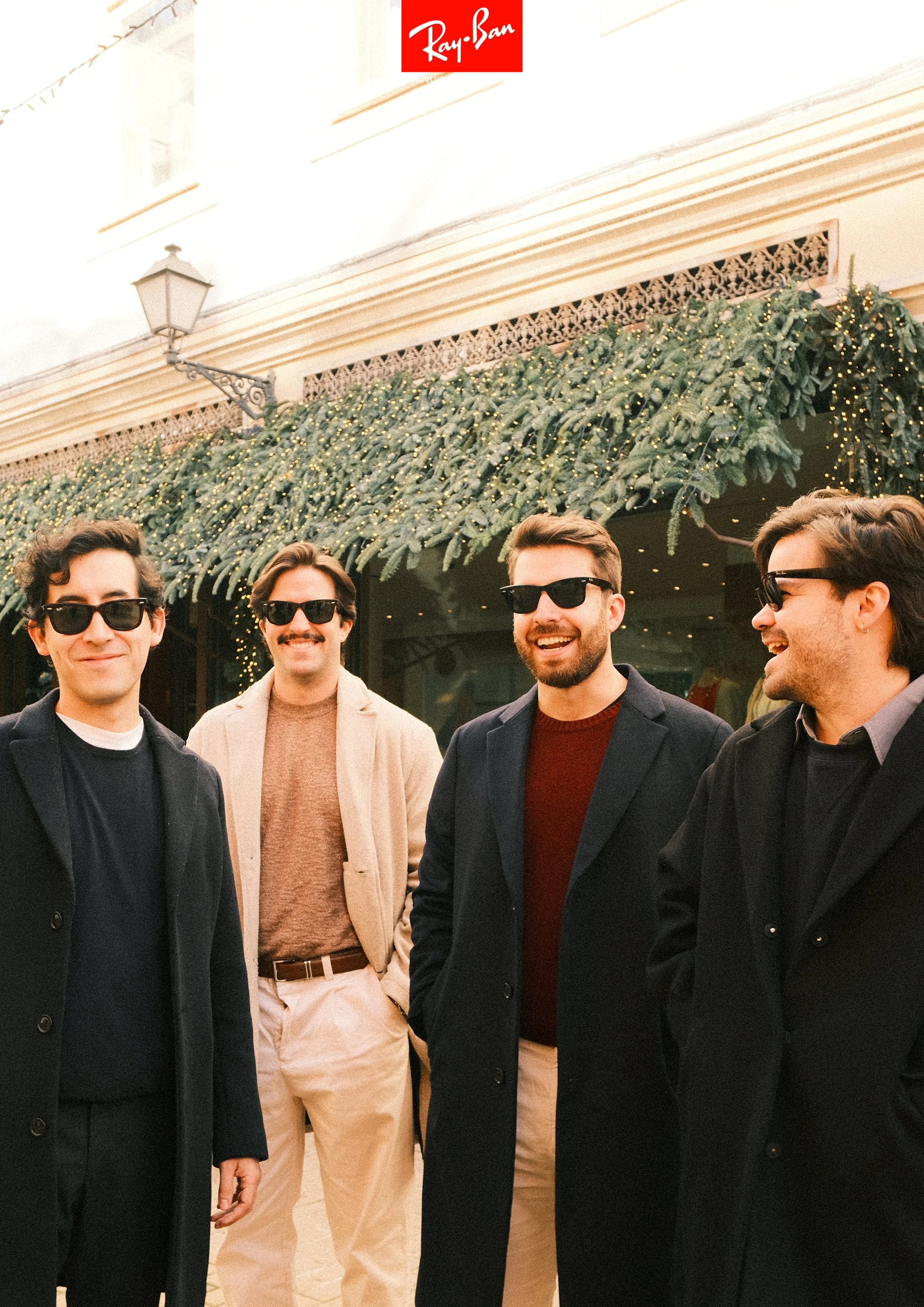 Four men wearing sunglasses and winter coats standing outside a decorated building with a Ray-Ban logo at the top, Christmas greenery, and string lights.