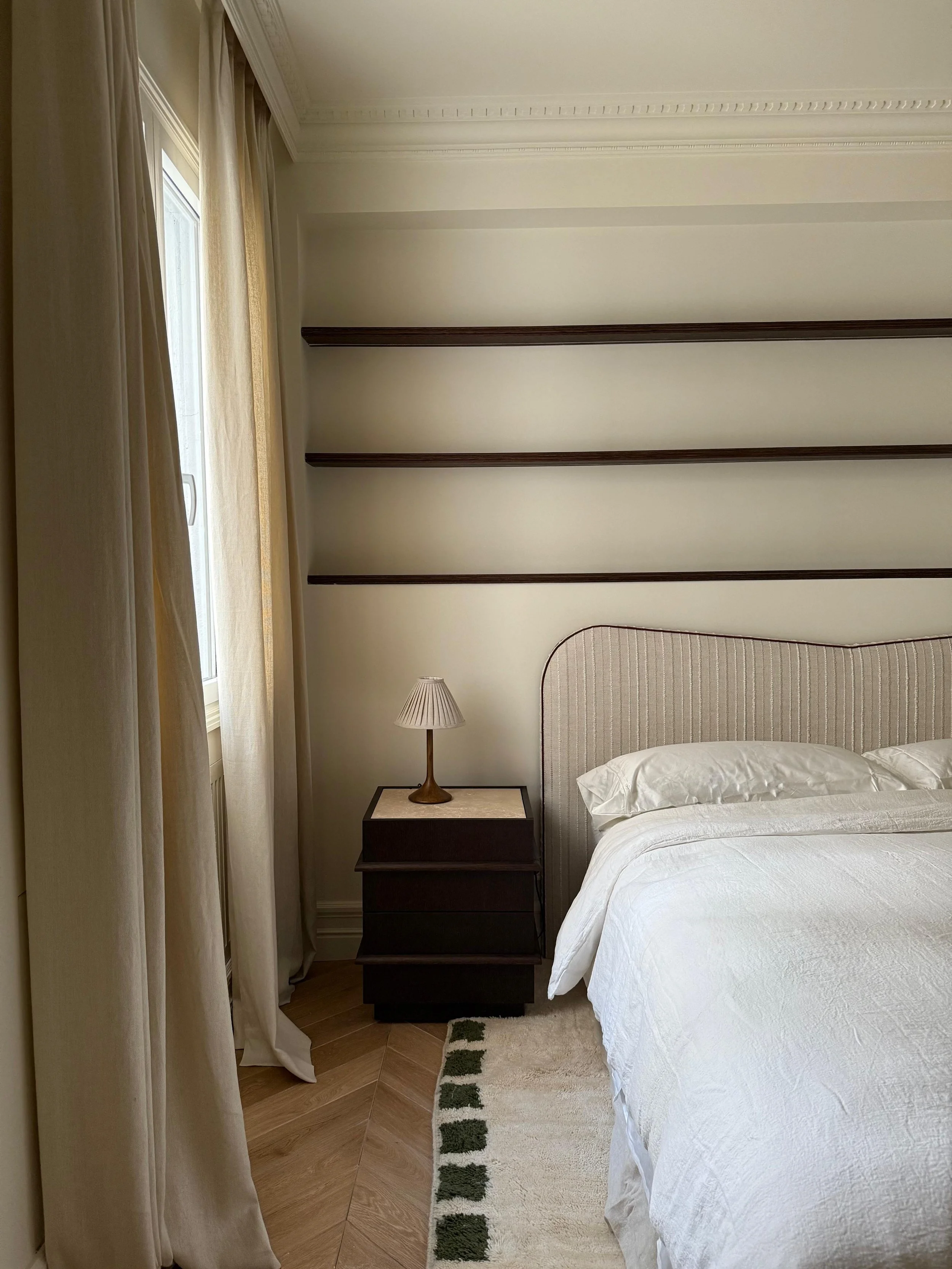A bedroom corner with a bed, nightstand with a lamp, window with curtains, and empty wall shelves.