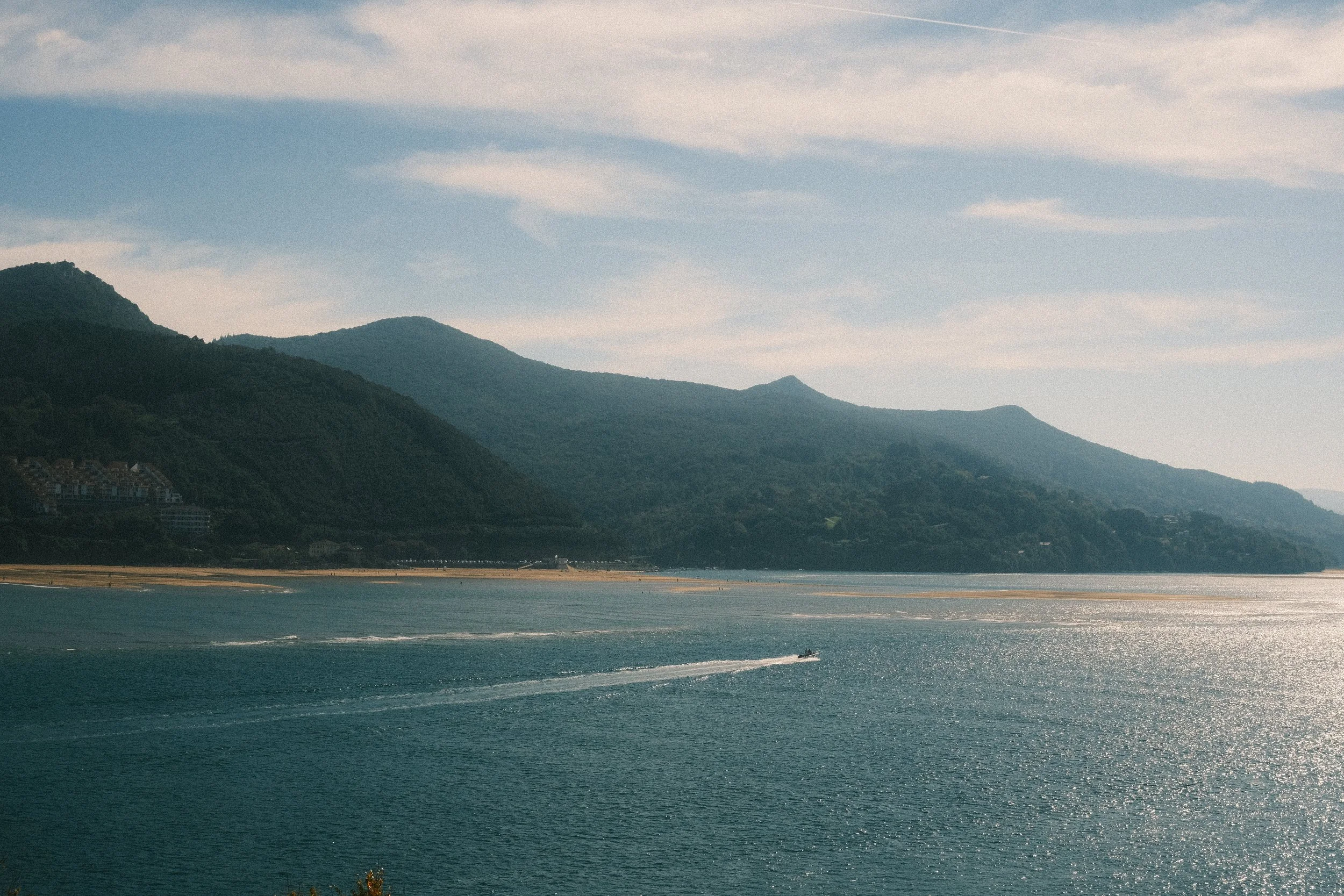 Scenic view of a mountain range along the coast with a body of water in the foreground, a small boat leaving a wake, and partly cloudy sky.