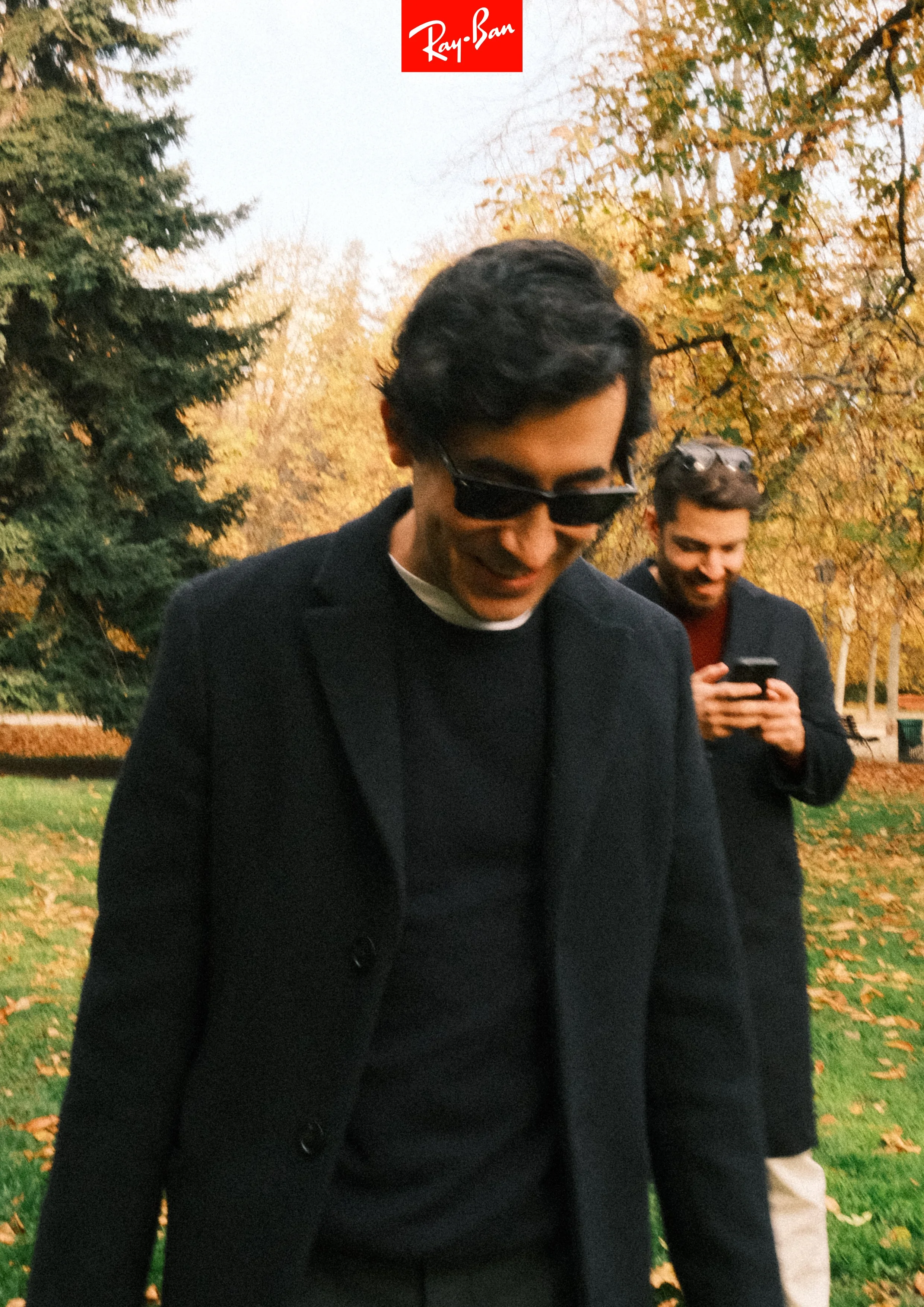 Two young men walking outdoors in autumn, one wearing sunglasses and a black coat, the other looking at a smartphone, with trees and fall foliage in the background.