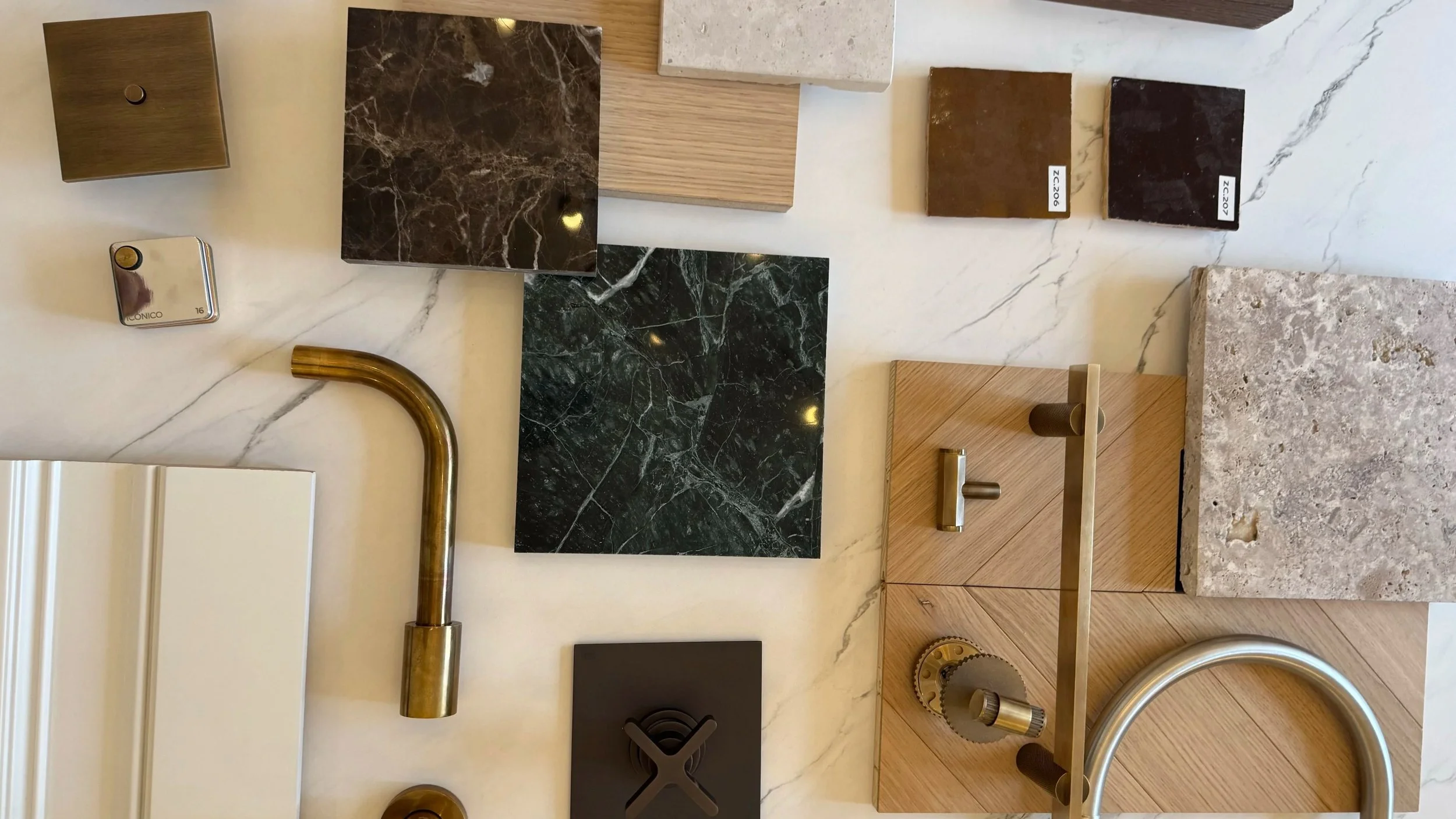 Collection of material sample tiles and fixtures including various marble, stone, wood, and metal finishes, along with a brass faucet and a kitchen handle on a white marble surface.